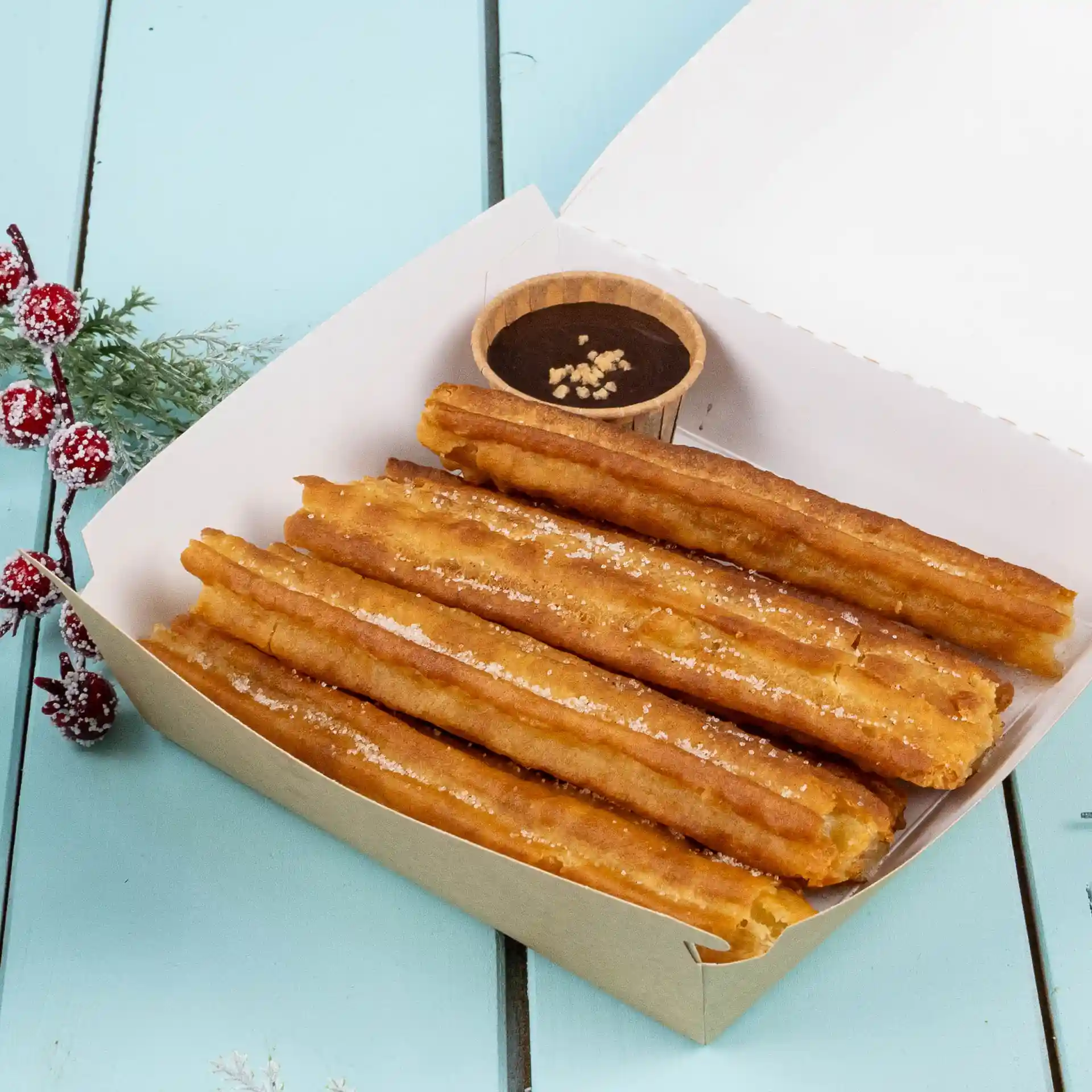 Churros w papierowym pudełku z małą brązową filiżanką sosu czekoladowego na niebieskiej powierzchni.