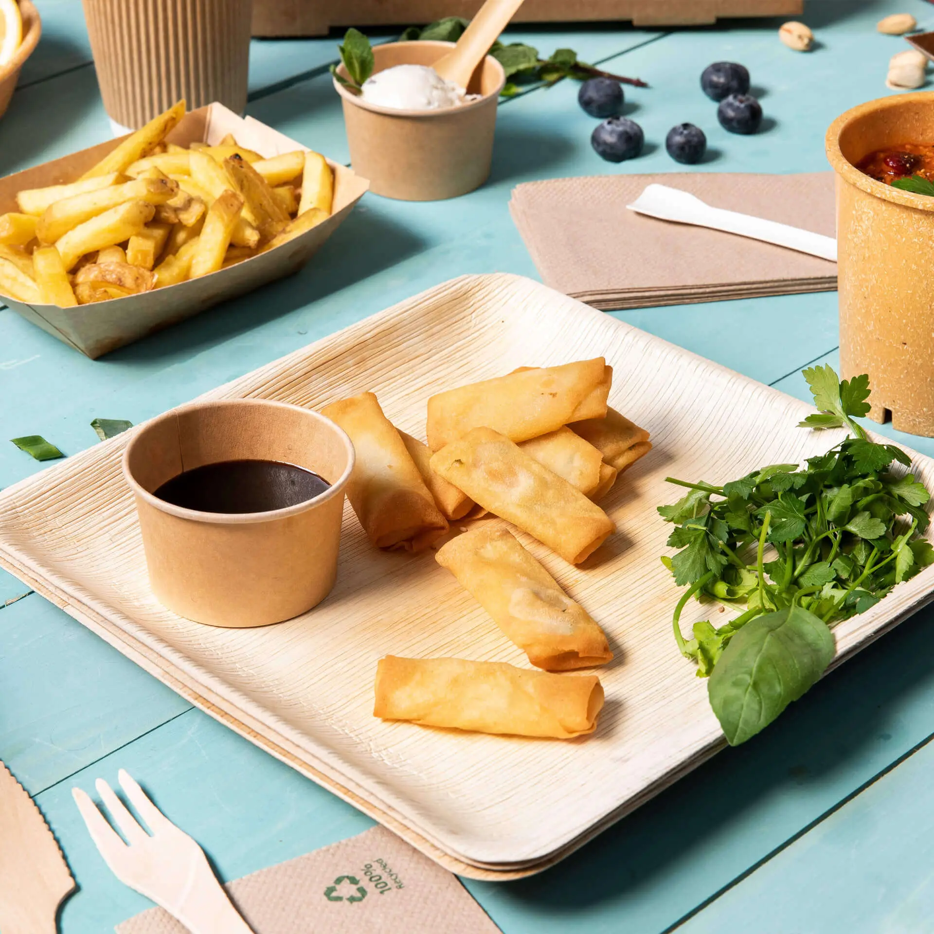 Sześć spring rolls na kwadratowym talerzu z liści palmowych; w towarzystwie małego kubka sosu do maczania i świeżych ziół.