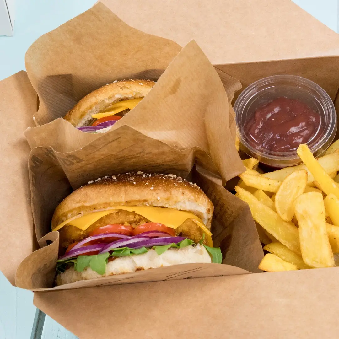 Dwa burgery z serem, sałatą, pomidorem i cebulą w pudełku na wynos, w towarzystwie frytek i małego pojemnika ketchupu.