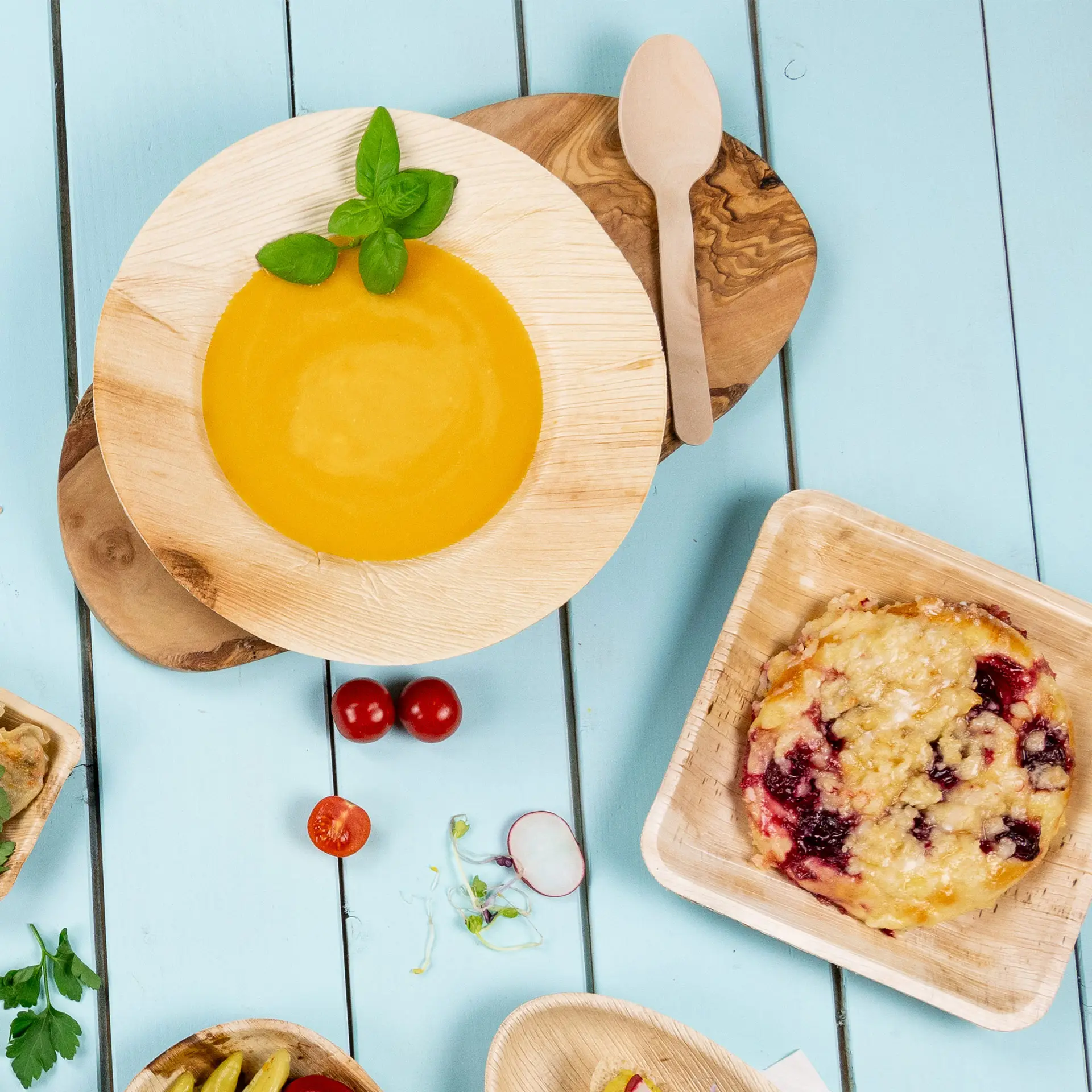 Jedna głęboka okrągła talerz z liścia palmowego z żółtą zupą i liśćmi bazylii na drewnianej desce; dodatkowe artykuły spożywcze i drewniana łyżka w pobliżu.