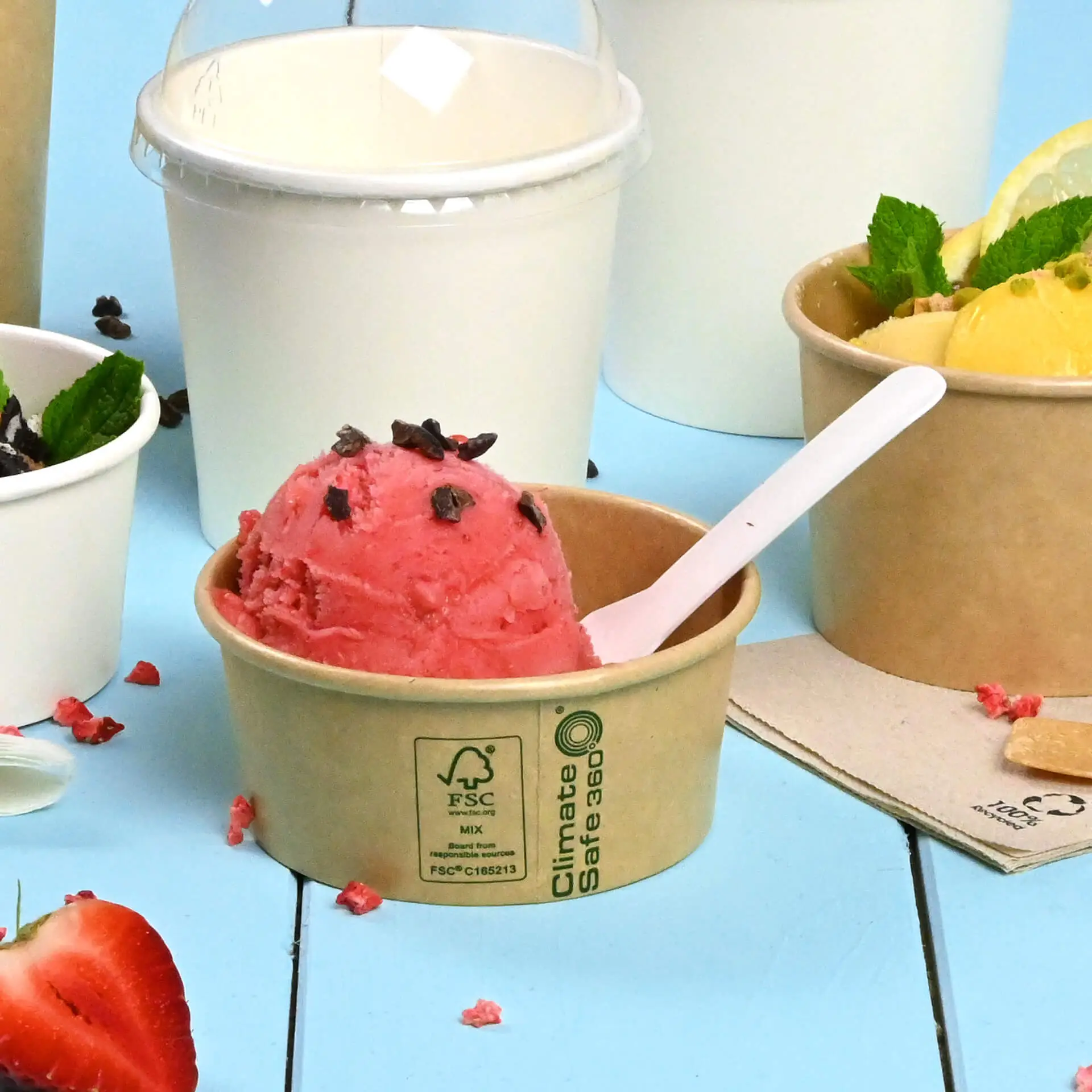 Brąski papierowy kubek lodów z różowym lodem truskawkowym i czekoladowymi posypkami, otoczony innymi pojemnikami na lody.