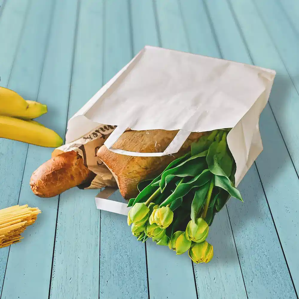 Biały papierowy worek wypełniony zakupami, w tym chlebem, tulipanami i bananami na niebieskiej drewnianej powierzchni.