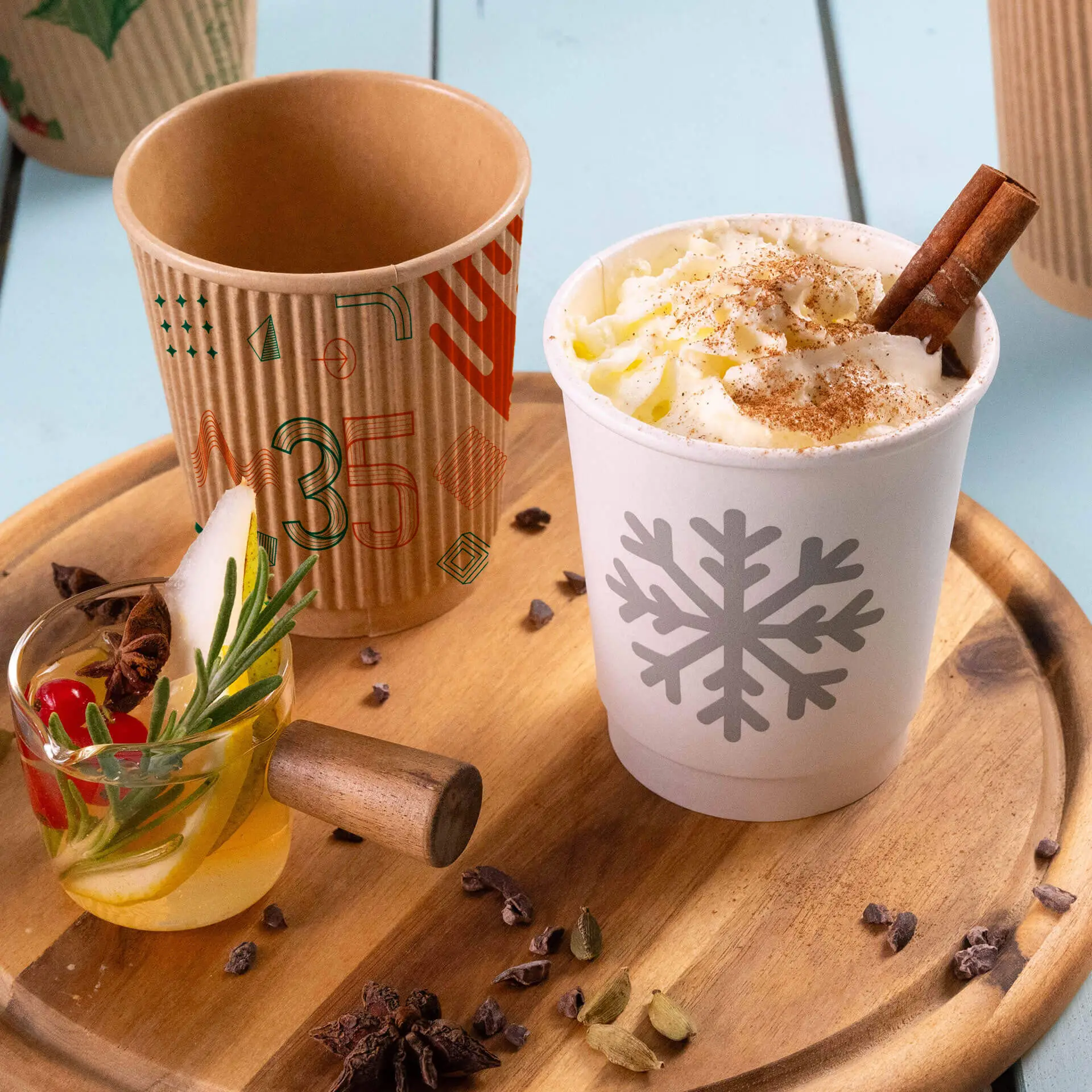 Jedna biała papierowa filiżanka z wzorem płatków śniegu, wypełniona bitą śmietaną i cynamonowym patyczkiem; obok brązowy prążkowany kubek.