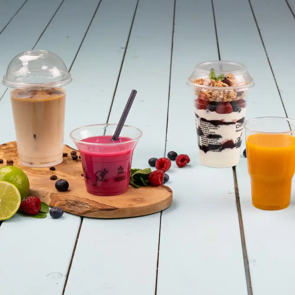 Cztery napoje na drewnianej desce: iced coffee w kubku z kopułą, różowy napój ze słomką, deser w kubku i sok pomarańczowy.