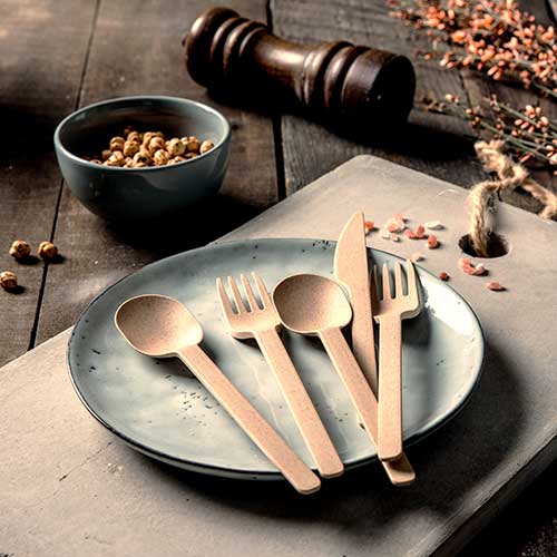 Biodegradowalny zestaw sztućców wielokrotnego użytku z łyżką, widelcem, nożem i talerzem na drewnianej powierzchni z miską przekąsek.