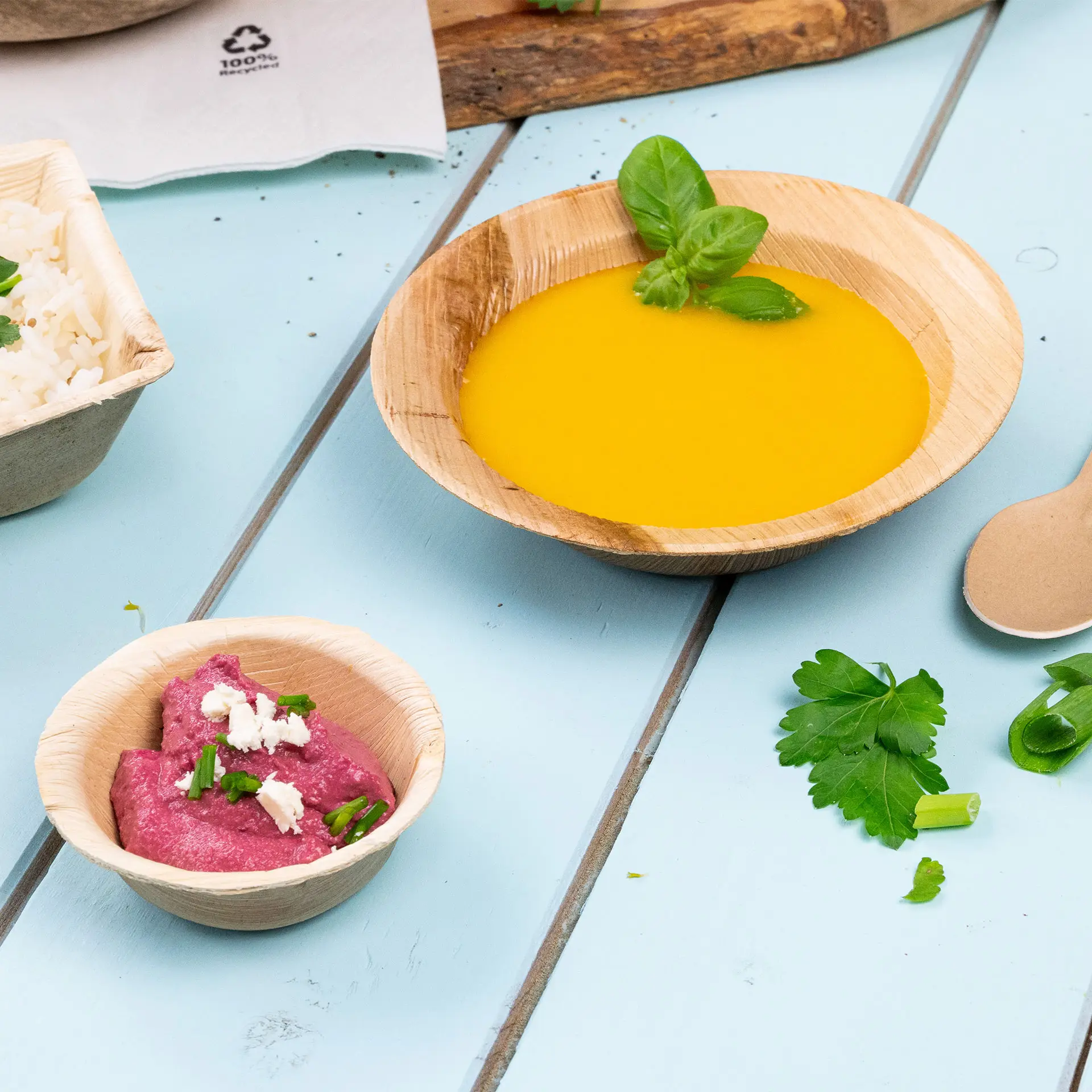 Okrągła mini miseczka z liścia palmowego z pomarańczowym sosem, udekorowana liściem bazylii; obok mniejsza miseczka z różowym dipem i ziołami.