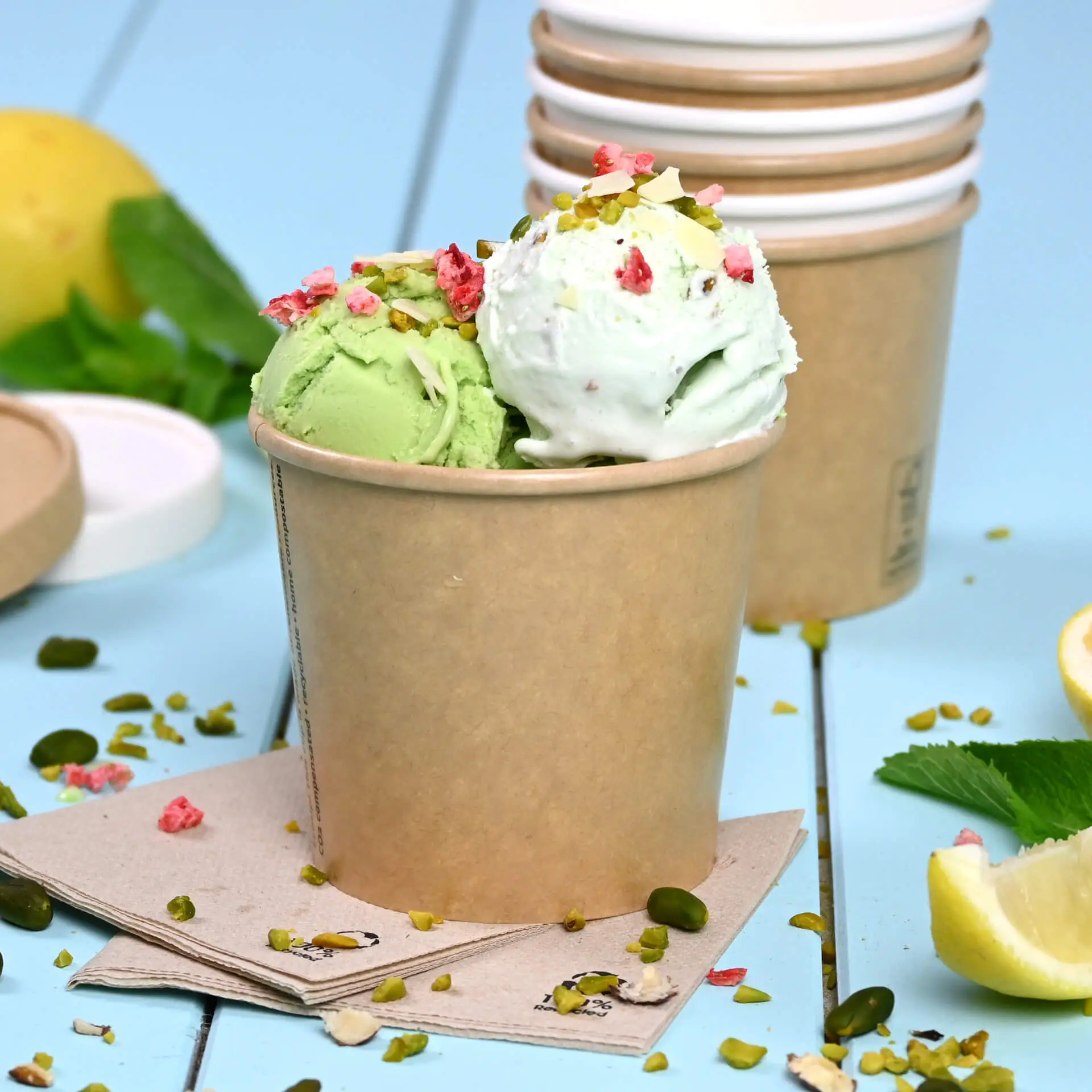 Brąski papierowy kubek na lody wypełniony zielonym i białym lodem, posypany orzechami i owocowym posypaniem; więcej kubków w tle.