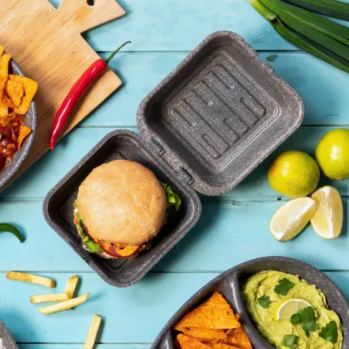 Burger w wielorazowej pudełku z kraftu z sałatą i sosami, otoczony chipsami, frytkami, guacamole i kawałkami limonki.