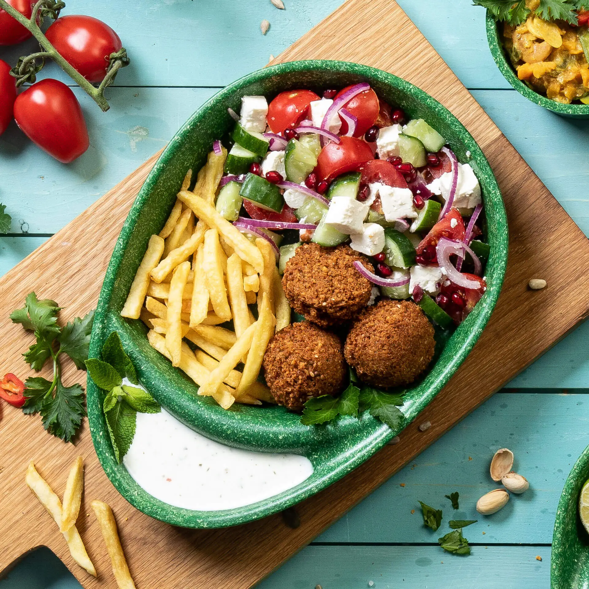 Wielokrotna podzielona taca w kolorze szpinaku z dwoma przegródkami; zawiera falafel, sałatkę, frytki i sos na drewnianej powierzchni.