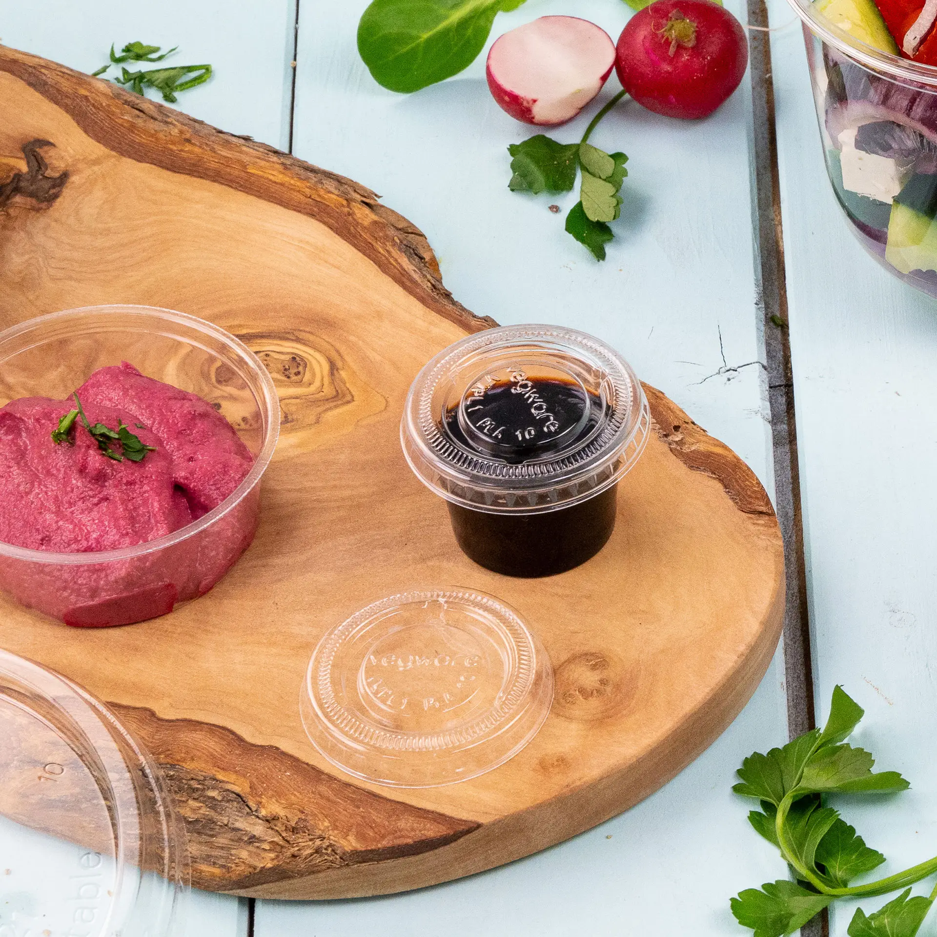 Jedna przezroczysta pokrywka i dwa małe pojemniki: jeden z różowym dipem, drugi z ciemnym sosem, na drewnianej powierzchni.