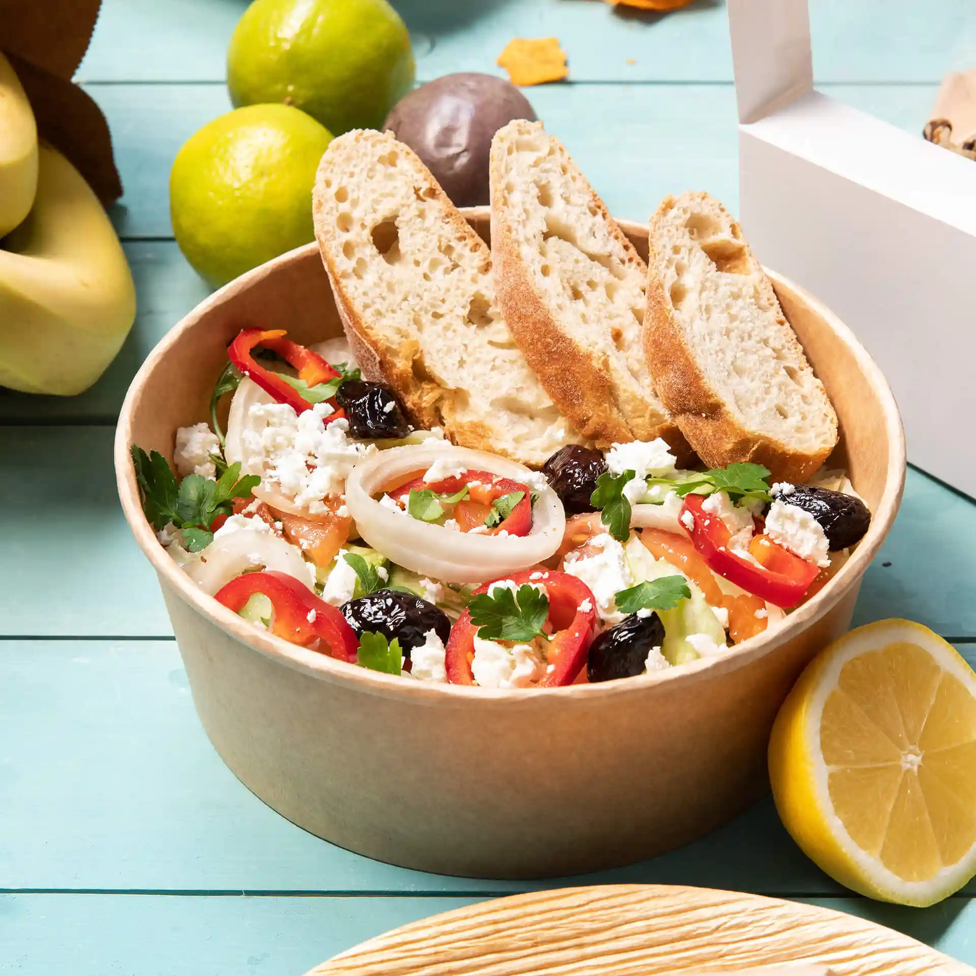 Miska wypełniona sałatką, z pokrojonymi warzywami i serem feta, w towarzystwie dwóch kromek chleba, na niebieskiej powierzchni.