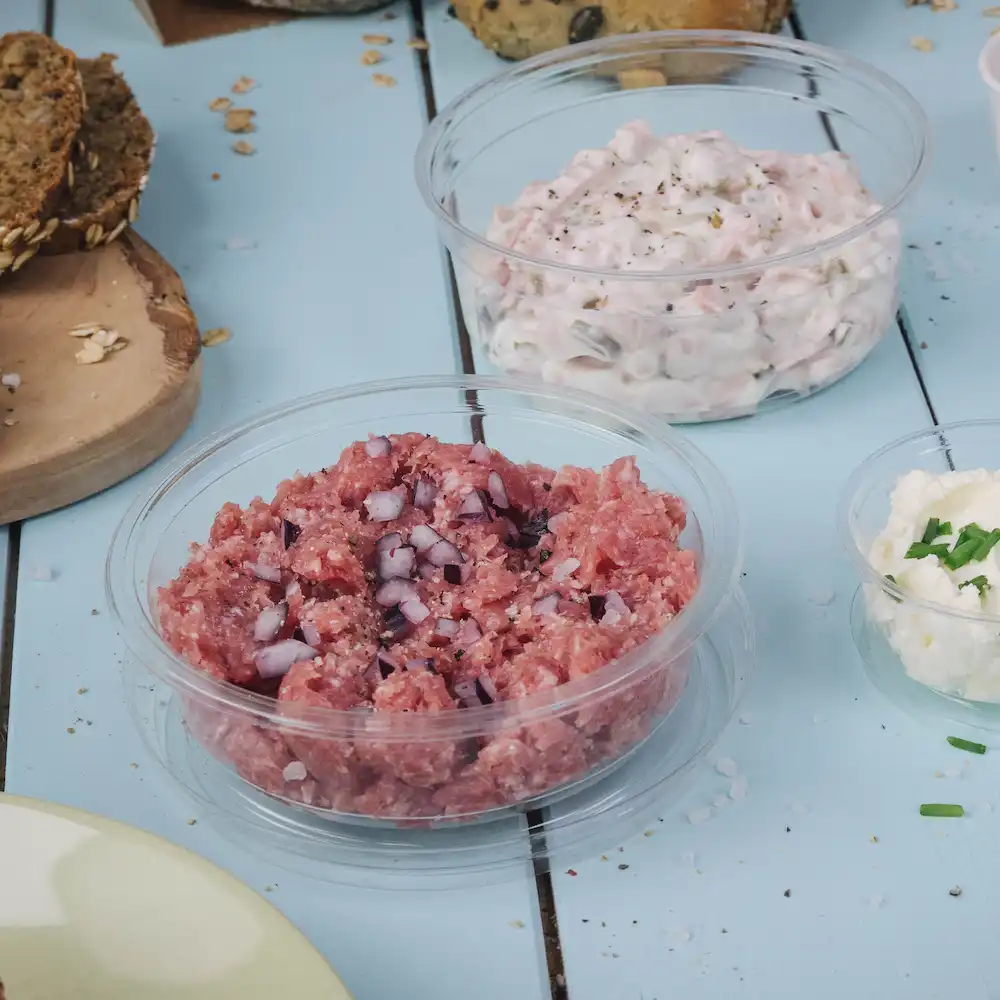 Siekanego surowego mięsa z pokrojoną cebulą w przezroczystym pojemniku; obok kremowy dip w innym pojemniku oraz mała miseczka sera.