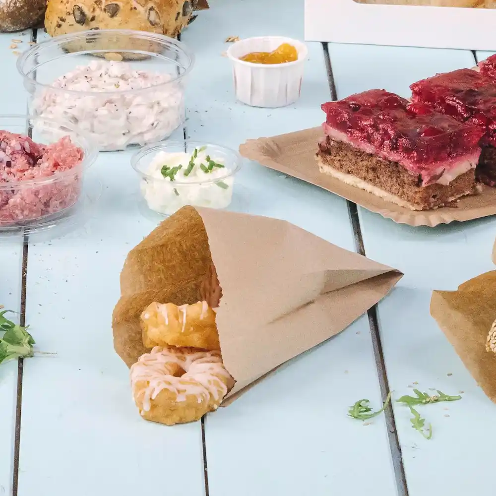 Brązka papierowa torba z dwoma lodowymi pączkami, otoczona różnymi produktami spożywczymi i dodatkami na niebieskim stole.