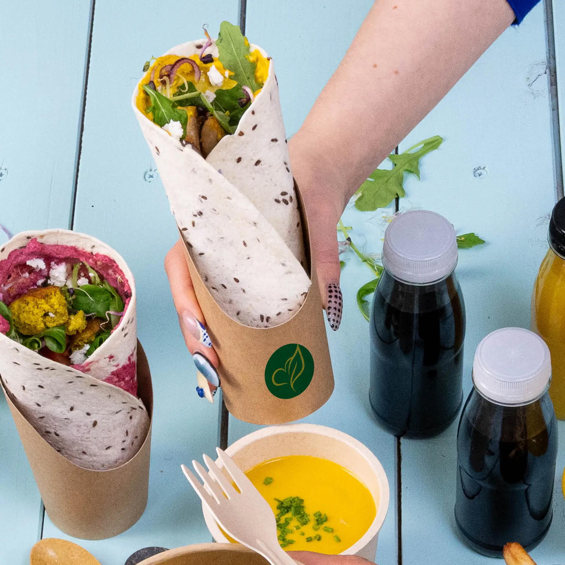 Jedna brązowa, spersonalizowana filiżanka wrap z wypełnionym wrapem; obok druga filiżanka. W pobliżu znajduje się kilka butelek i miska z sosem.