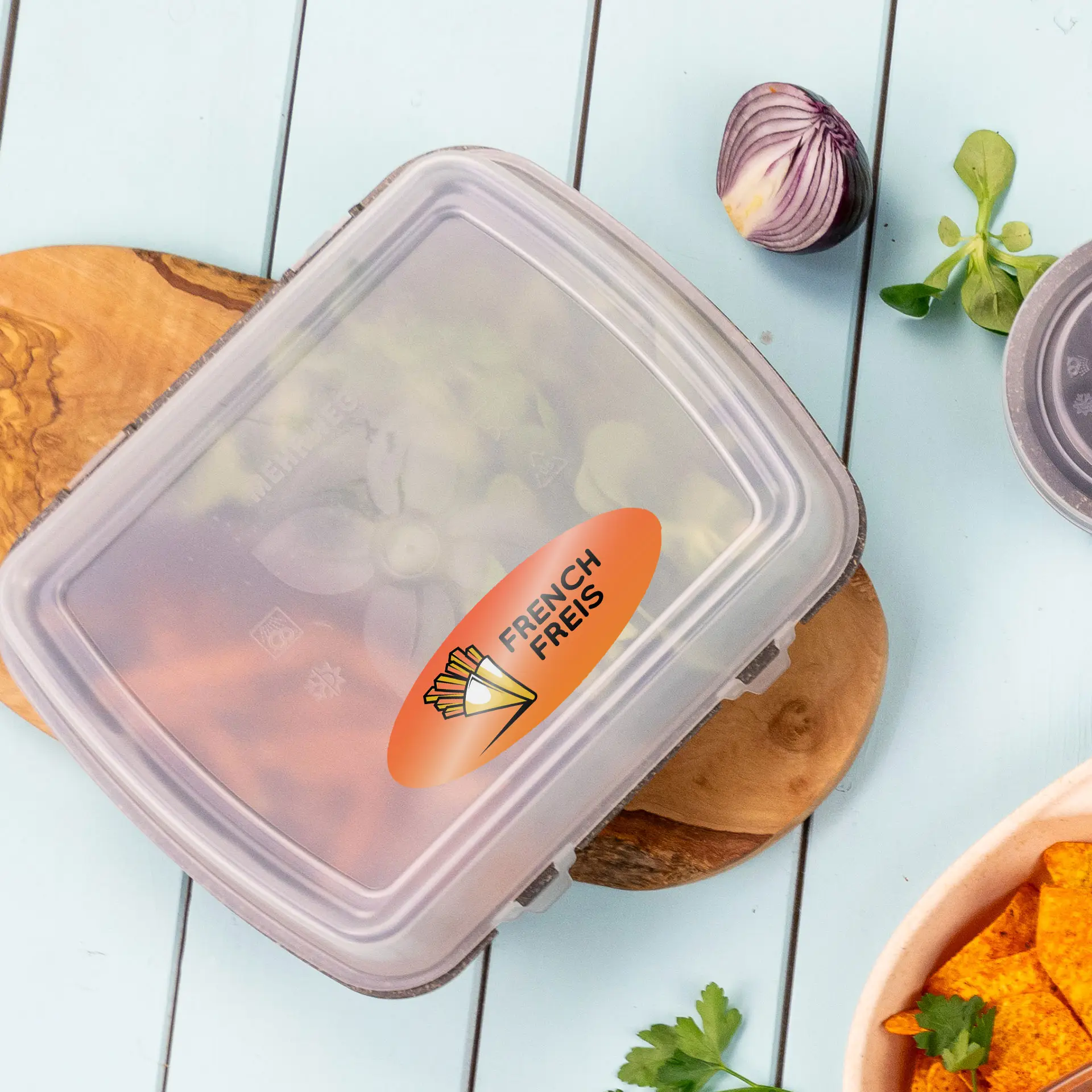 Przezroczysta owalna etykieta z napisem "FRENCH FRIES" na pojemniku, otoczona świeżymi składnikami na drewnianej desce.