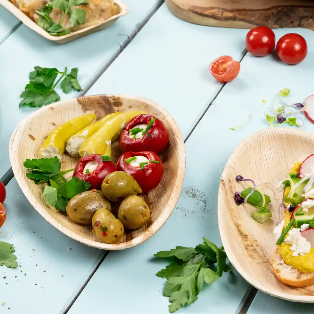 Drewniana deska z nadziewanymi czerwonymi paprykami, marynowanymi żółtymi paprykami i zielonymi oliwkami; świeże zioła rozsypane na niebieskiej powierzchni.