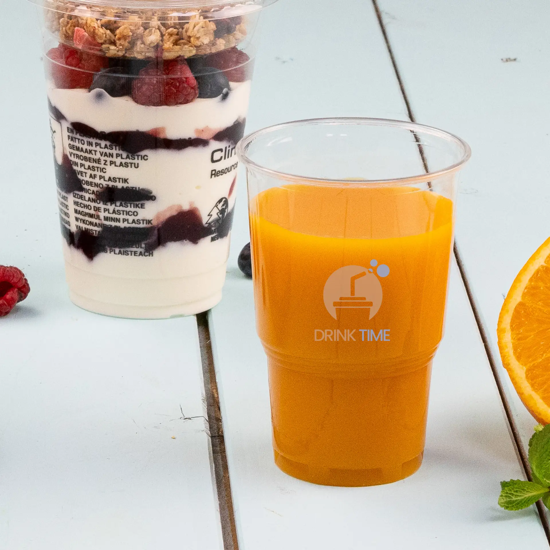 Przezroczysty kubek PLA 10 oz z pomarańczowym napojem i grafiką diamentową; pokrojona pomarańcza i mięta obok; parfait w tle.