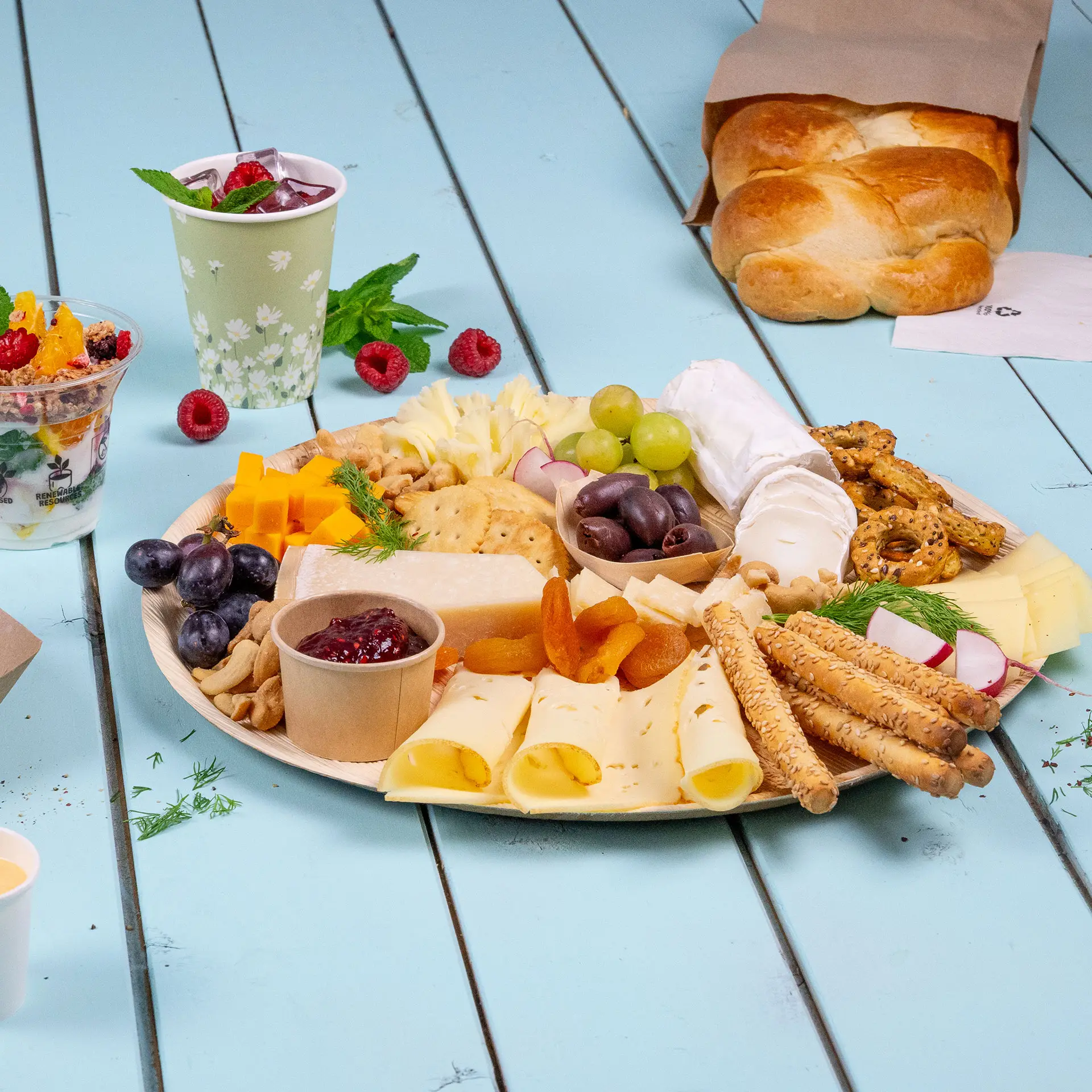 Okrągły talerz z liścia palmy z wyborem serów, owoców, orzechów i krakersów, a także bochenkiem chleba i napojami.