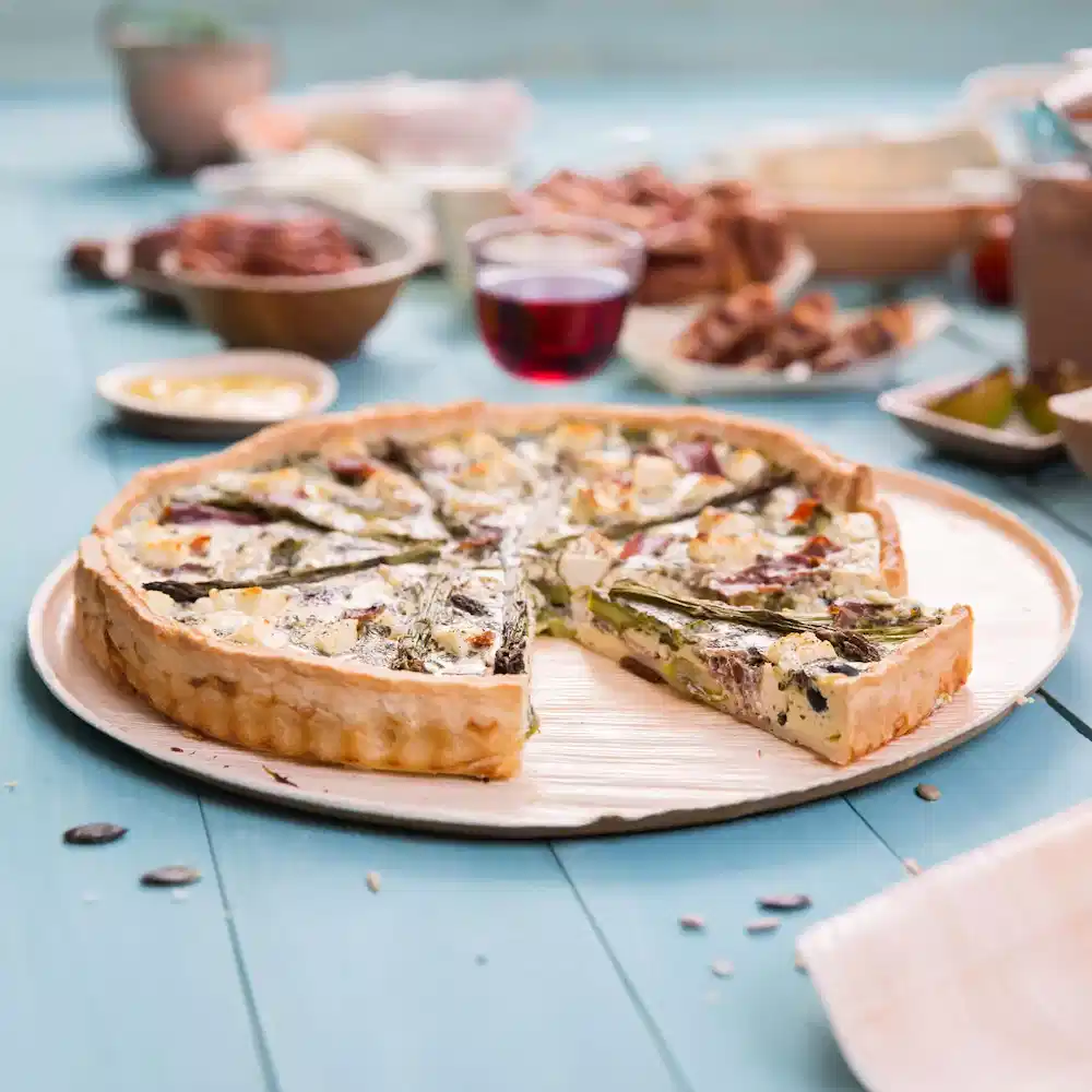 Słona quiche na drewnianym talerzu, z wyjętym kawałkiem, otoczona różnymi daniami i szklanką czerwonego napoju.