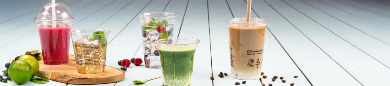 Różnorodne napoje wyeksponowane na drewnianej powierzchni, w tym iced coffee, smoothie, parfait jogurtowy i sok, z plasterkiem pomarańczy obok.