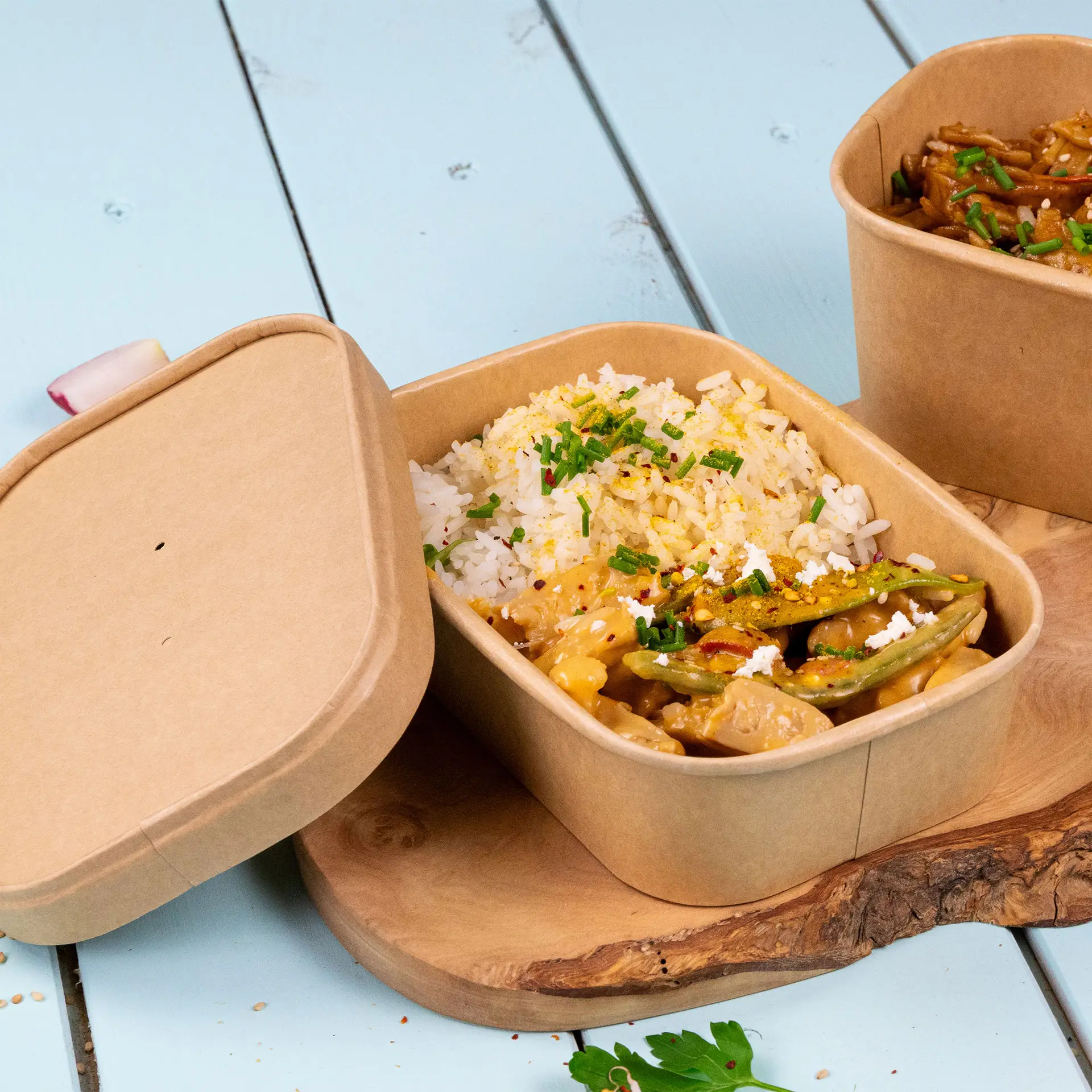 Prostokątny pojemnik kraft na jedzenie z ryżem i warzywami, lekko otwarty; pokazany na drewnianej desce z podobnym pojemnikiem obok.