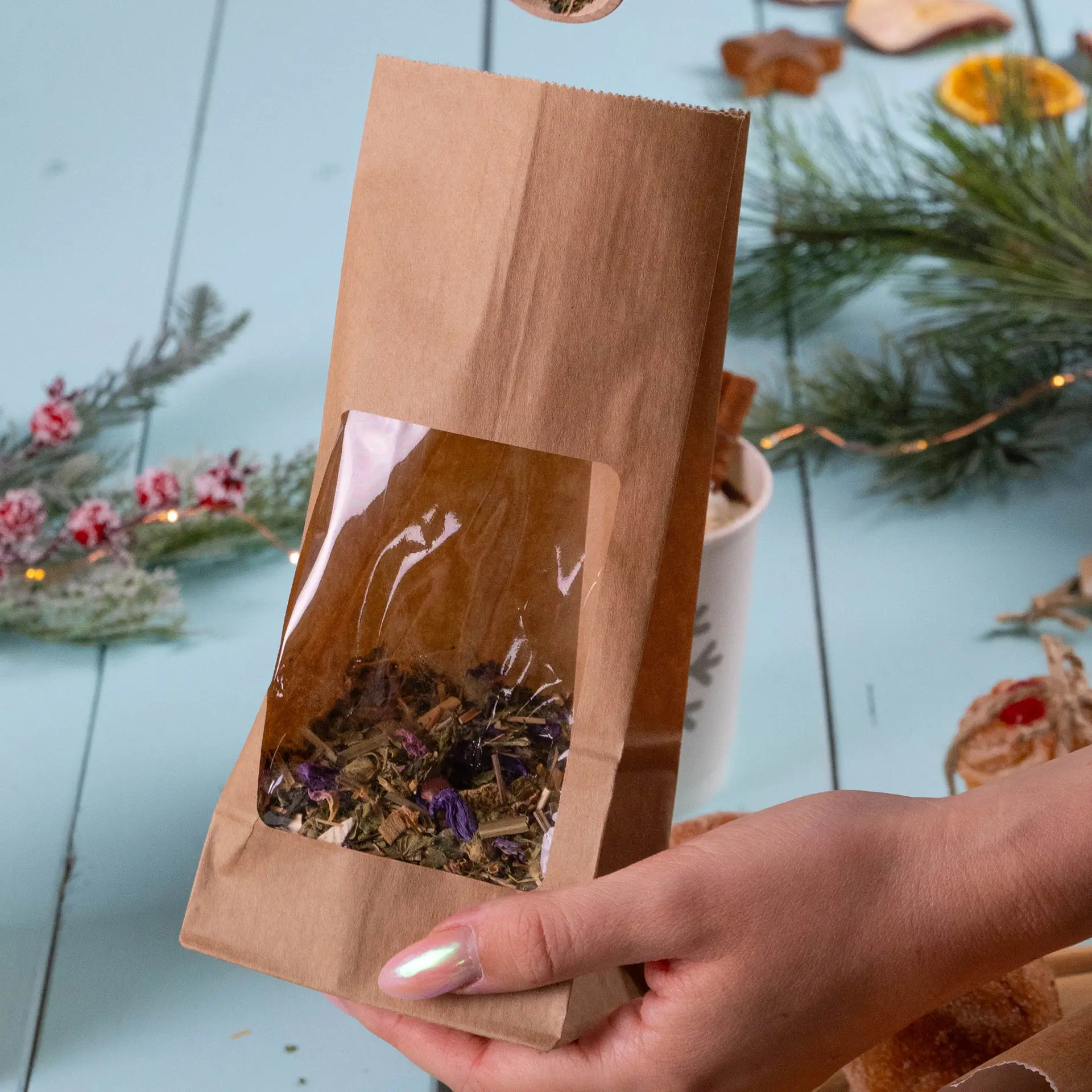 Ręka trzyma brązową torbę papierową z przezroczystym oknem, zawierającą luźne zioła; elementy dekoracyjne w tle.