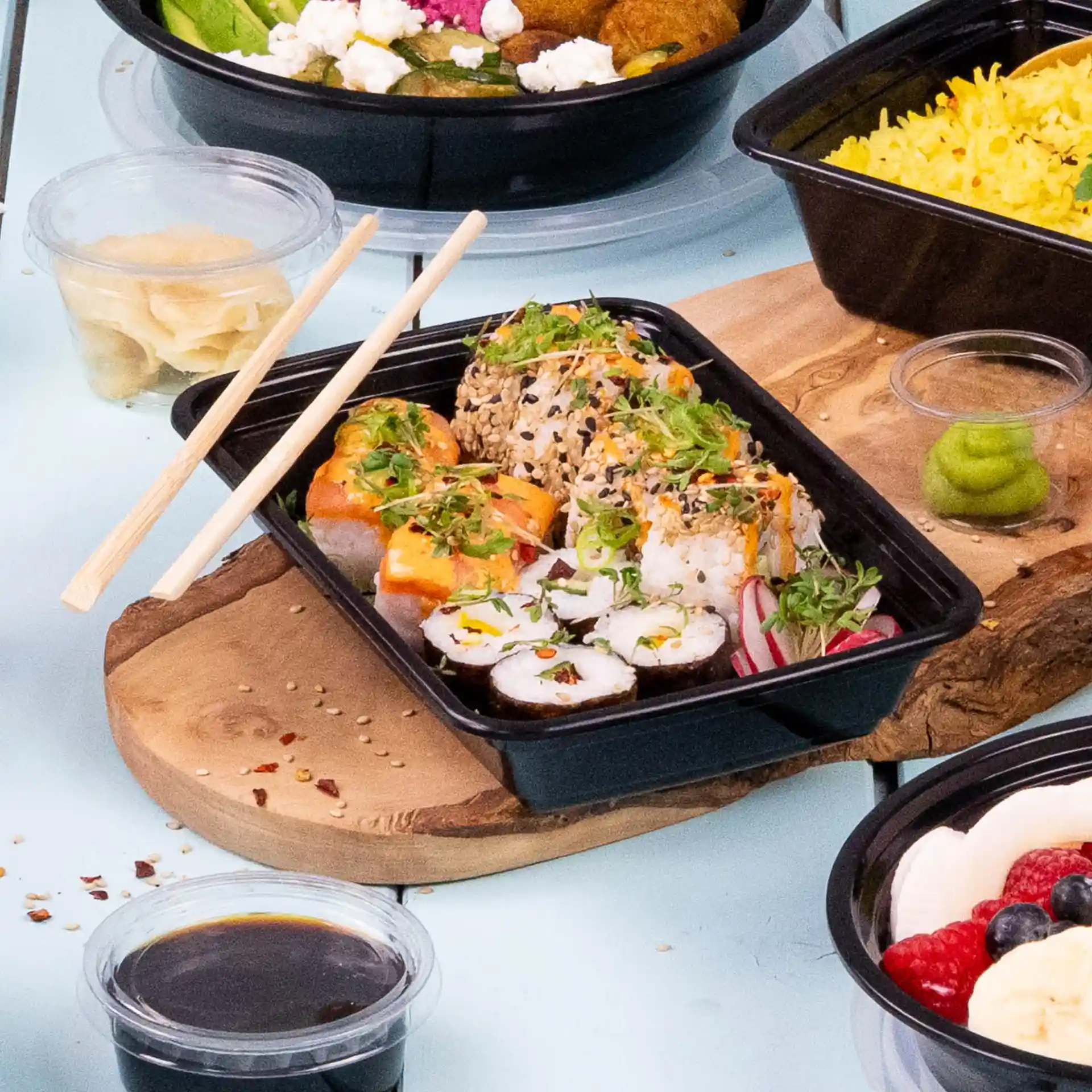 Rolki sushi z dodatkami w czarnym prostokątnym pojemniku wielokrotnego użytku, pałeczki obok i przyprawy, w tym wasabi i sos sojowy.