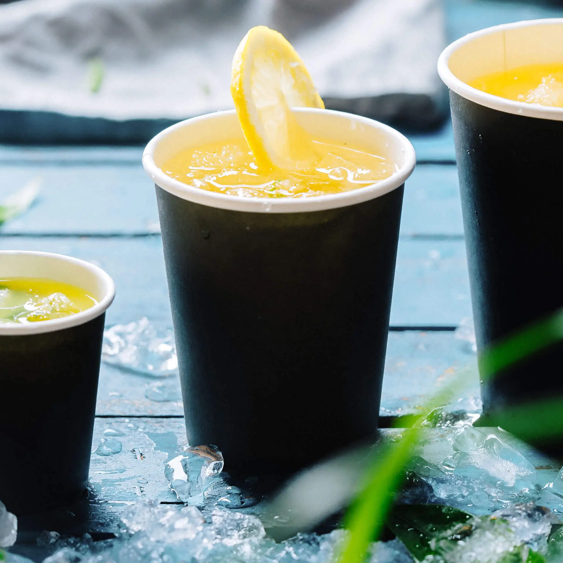 Dwa czarne papierowe kubki wypełnione napojami na drewnianym stole; lód i zieleń w tle. Większy kubek ma plasterek cytryny.