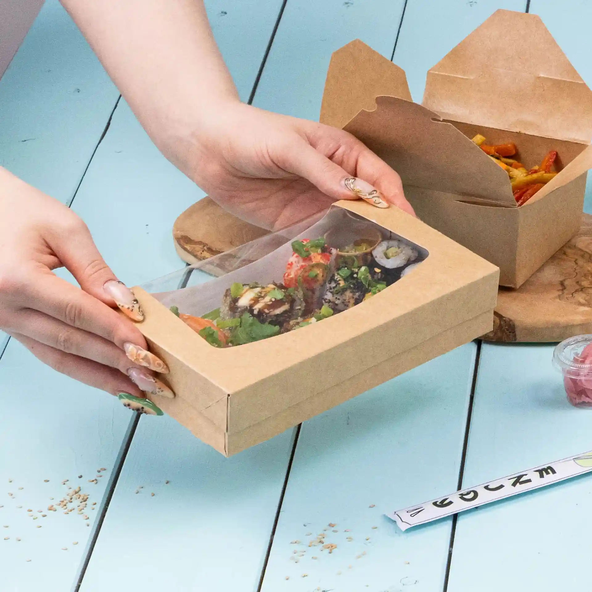 Kartonowe pudełko z sushi i sałatką, z przezroczystą górą, na niebieskiej powierzchni.