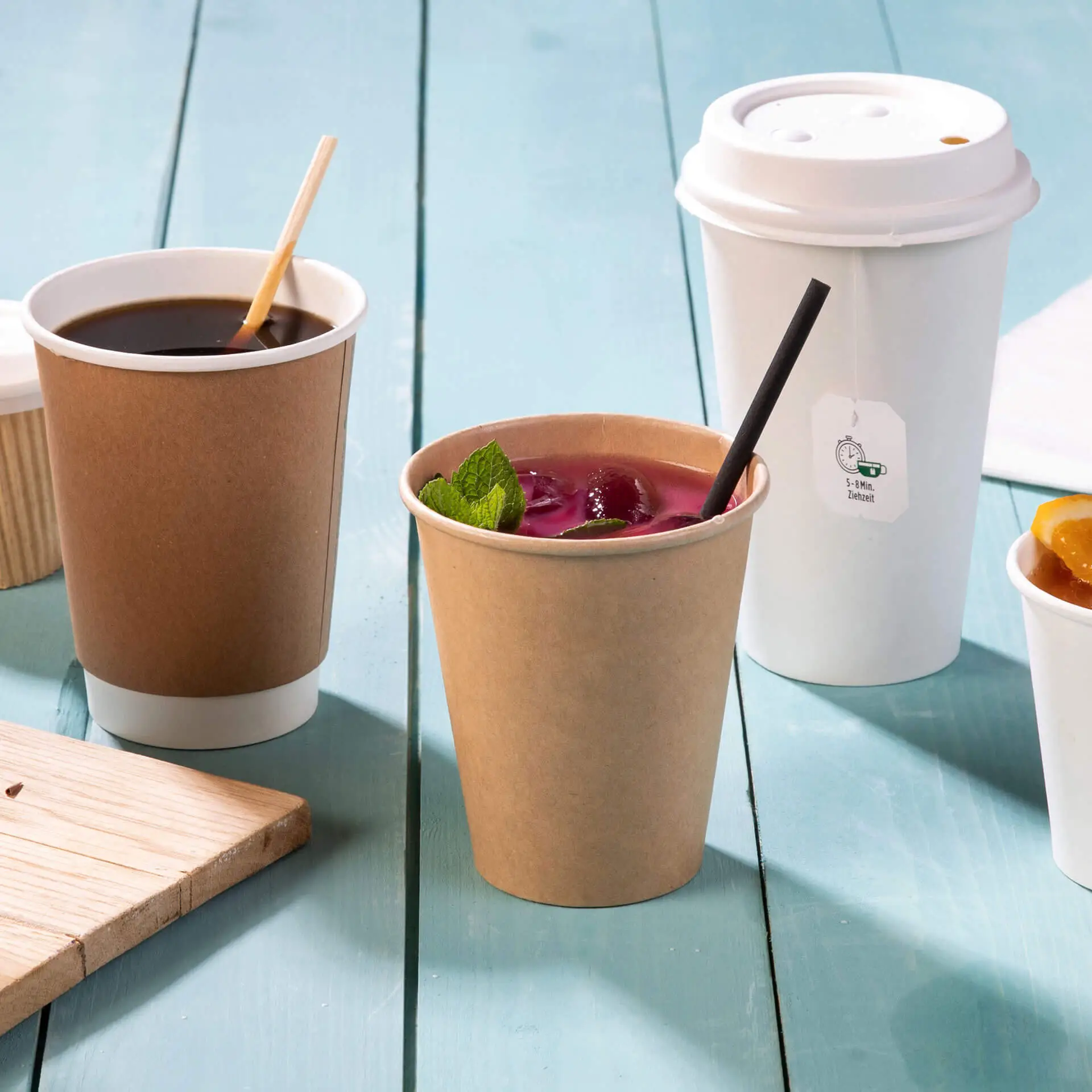 Brąski kubek papierowy ze słomką i kolorowym napojem, otoczony filiżanką kawy i innymi przedmiotami na jasnoniebieskim stole.