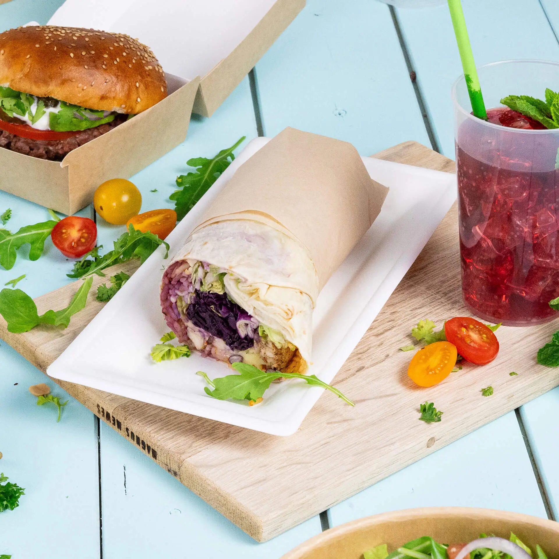 Prostokątna talerzyk z trzciny cukrowej z rolką wypełnioną warzywami, burgerem, pomidorkami koktajlowymi i napojem.