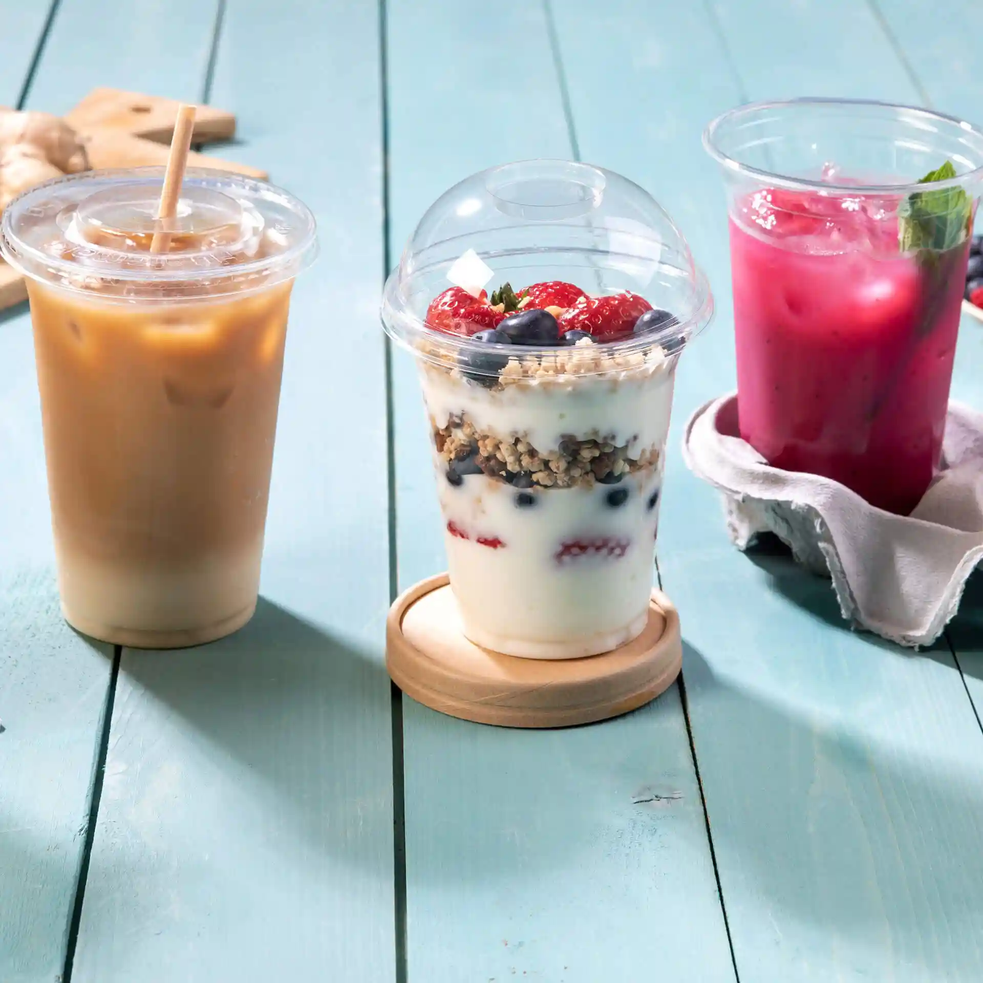 Obraz trzech przezroczystych plastikowych kubków na drewnianej powierzchni: iced coffee, jogurt z granolą i jagodami oraz różowy napój.