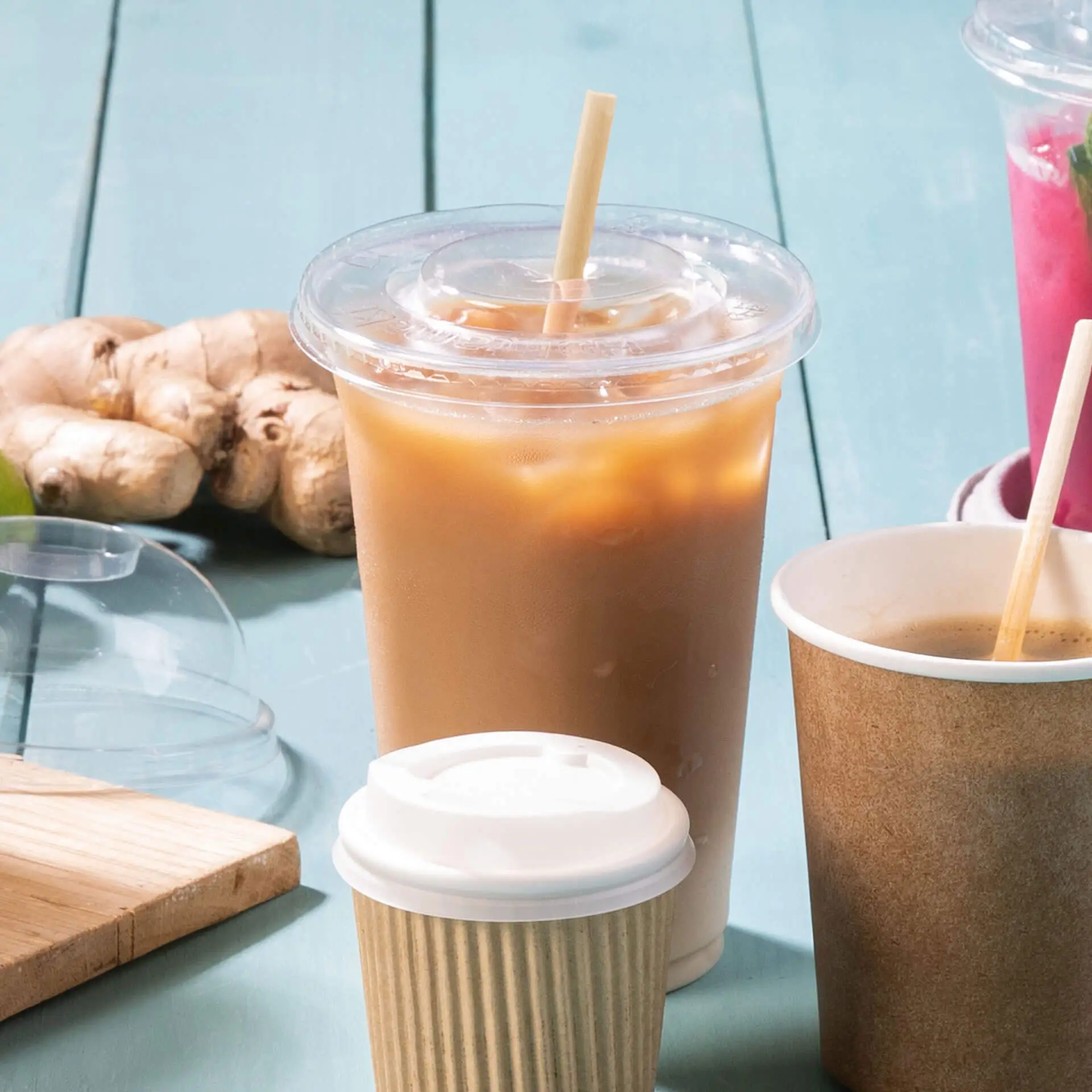 Jedna brązowa, biodegradowalna słomka papierowa z drewnianym patyczkiem, umieszczona obok napojów na lodzie i korzenia imbiru na tealowej powierzchni.