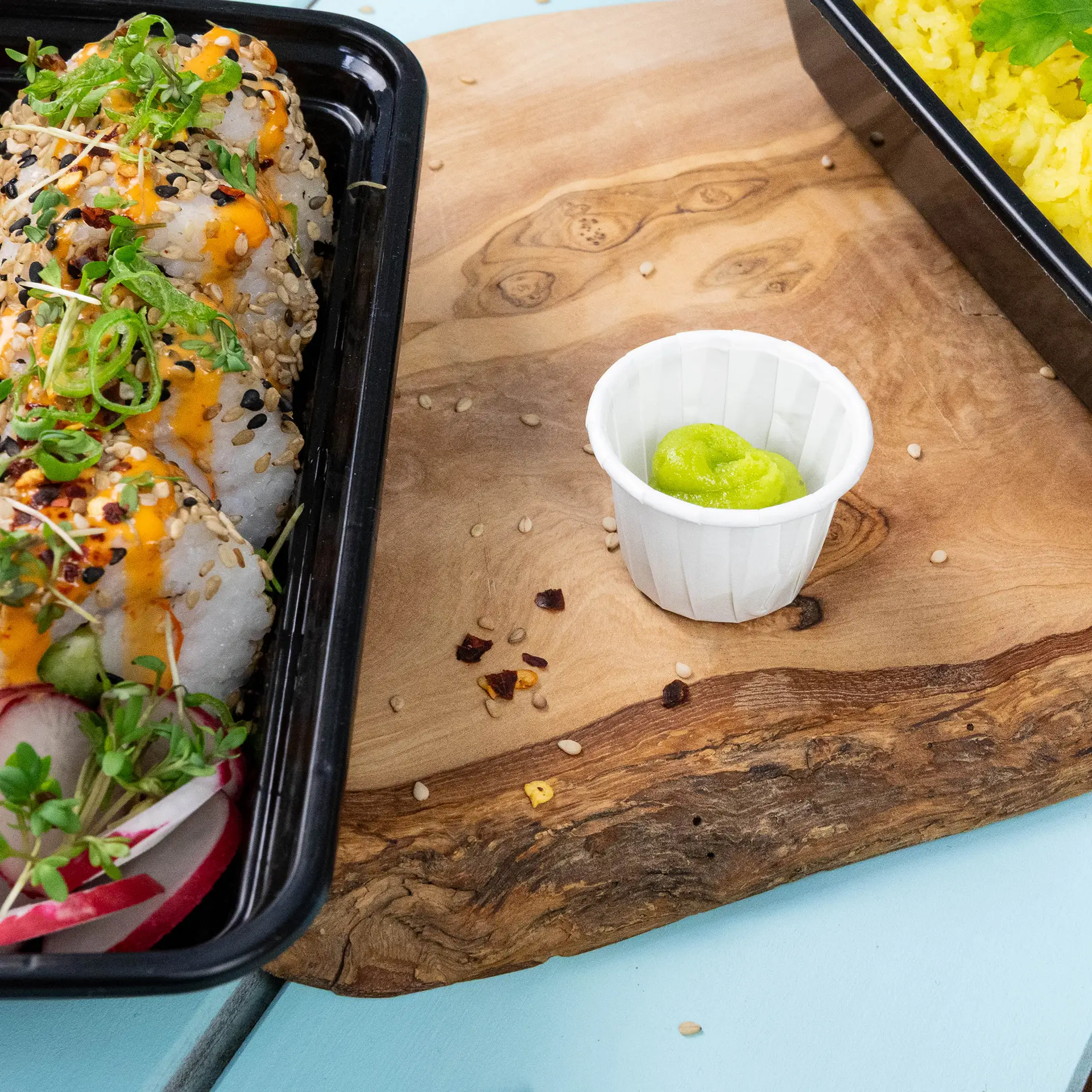 Czarny pojemnik z rolkami sushi, posypany sosem i sezamem, obok małego białego papierowego kubka z wasabi na drewnianej powierzchni.