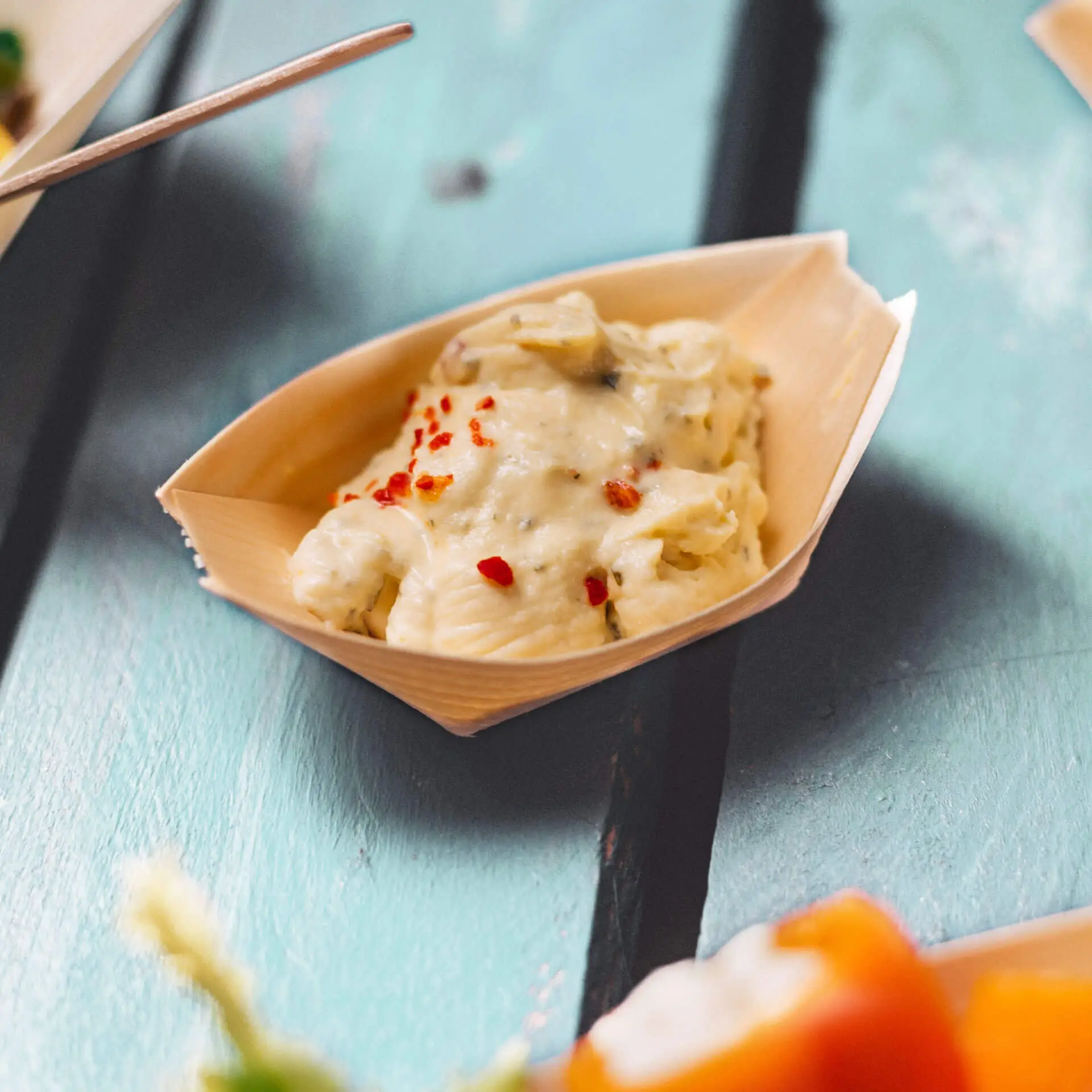 Drewniana miska w kształcie łodzi zawierająca kremowy posiłek posypany czerwonymi płatkami, na turkusowej drewnianej powierzchni.