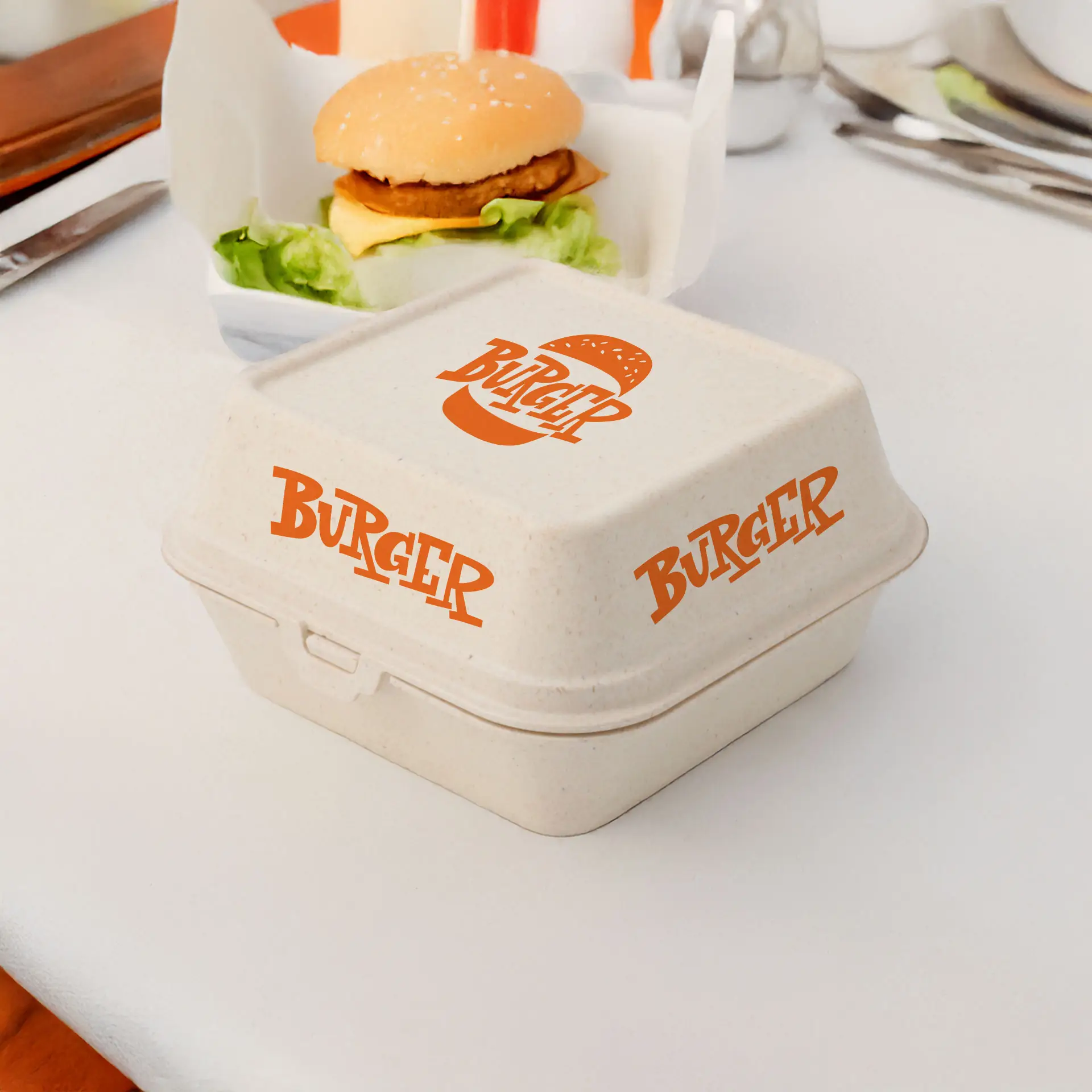 Kremowa prostokątna pudełko na burger z pomarańczowym nadrukiem, umieszczone na stole obok burgera i sałaty.