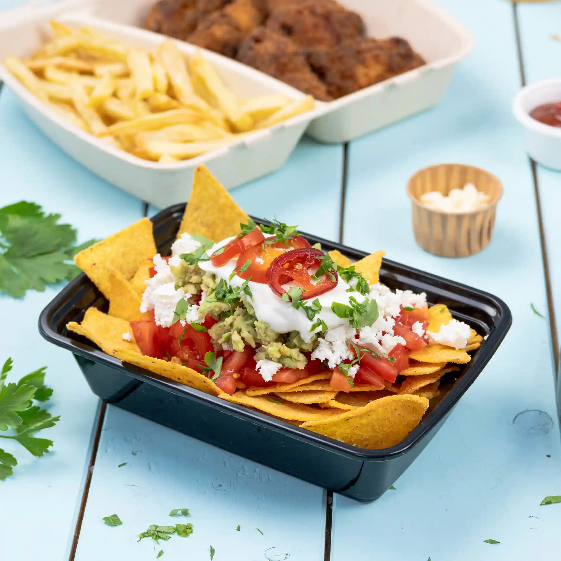 Czarny prostokątny pojemnik wielokrotnego użytku wypełniony nachos, z pomidorami, guacamole, śmietaną i kolendrą na wierzchu.