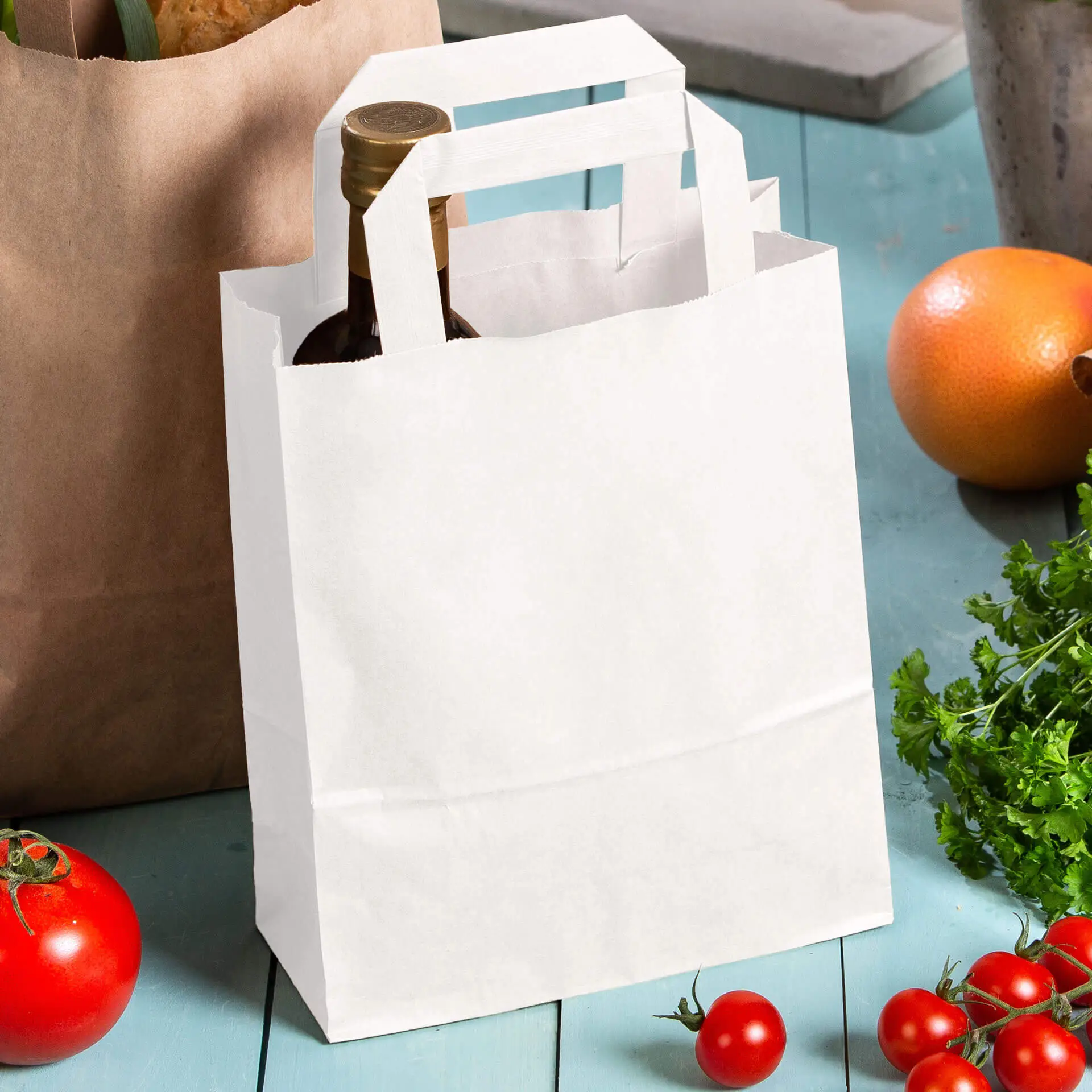 Biała torba papierowa z uchwytami, trzymająca butelkę, umieszczona obok pomidorów i innych zakupów.