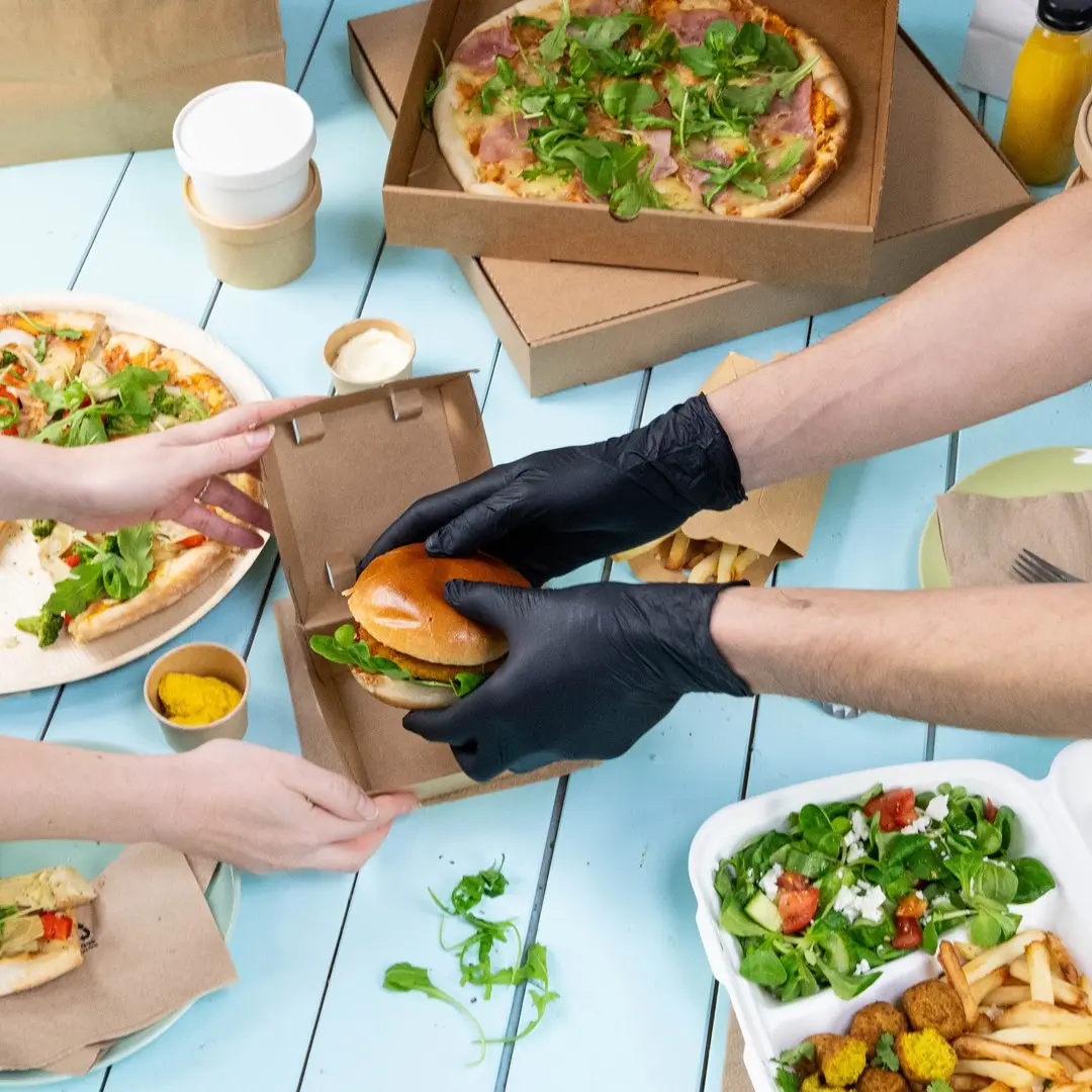 Ręce w czarnych rękawiczkach wkładają burgera do brązowego pudełka na wynos, otoczonego pizzą, sałatką i sosami.