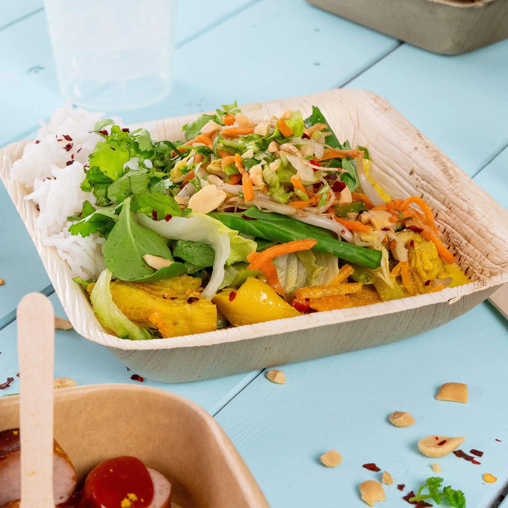 Miska z liści palmowych wypełniona ryżem, sałatką i warzywami, na jasnoniebieskiej powierzchni.