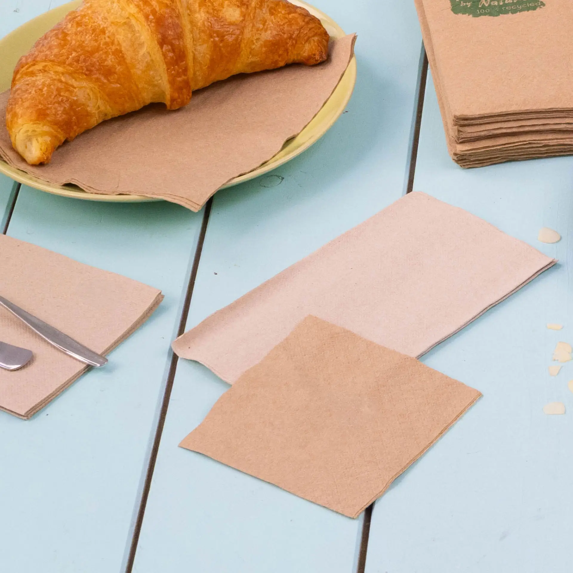 Obraz croissanta na żółtym talerzu z kilkoma brązowymi, recyklingowanymi serwetkami koktajlowymi na niebieskim stole.