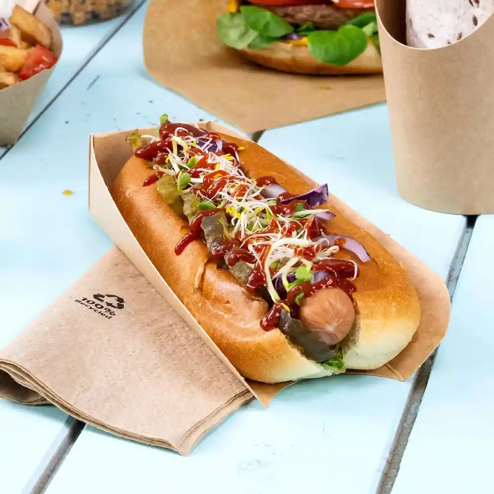 Hot dog w brązowym papierowym uchwycie, posypany ketchupem, musztardą, ogórkami i kiełkami, na drewnianym stole.