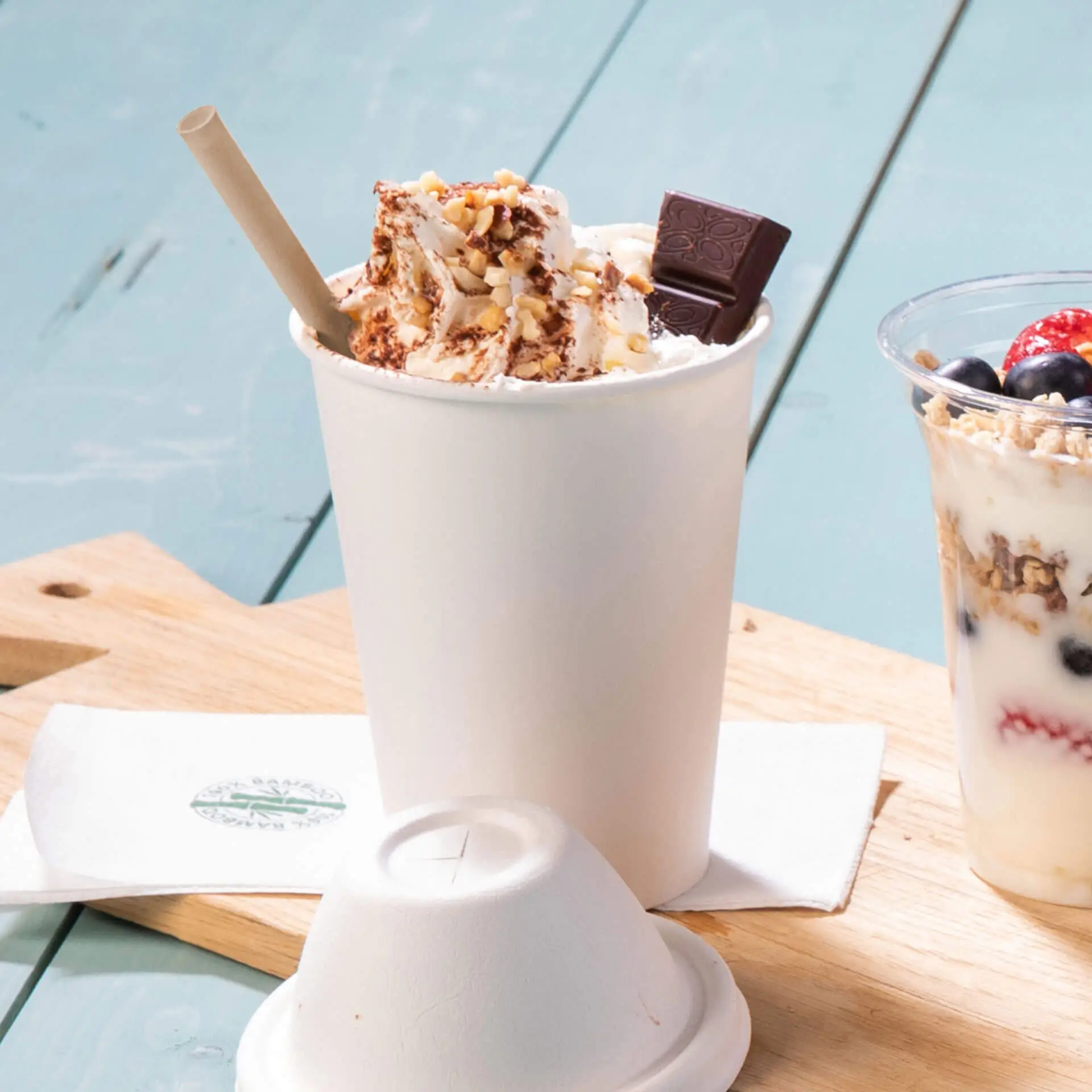 Biały papierowy kubek wypełniony bitą śmietaną, posypany orzechami i kawałkiem czekolady, obok przezroczystego kubka jogurtu parfait.