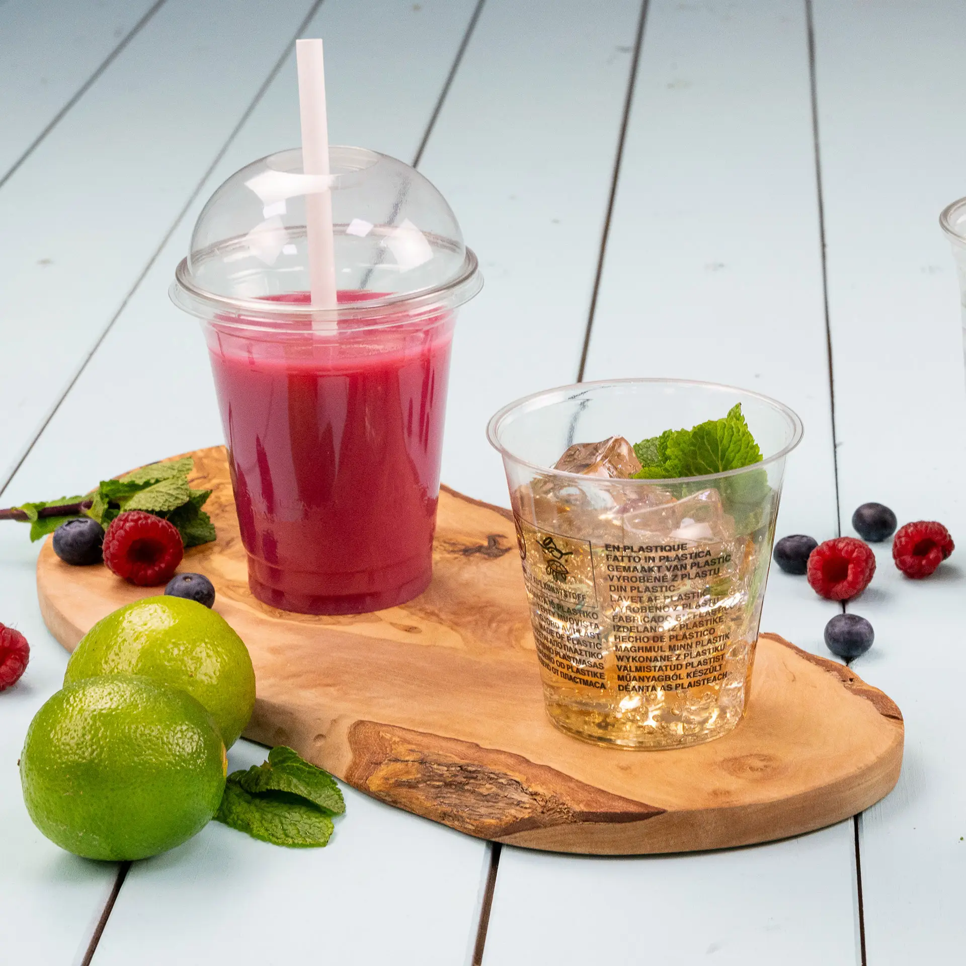 Dwa przezroczyste kubki rPET na drewnianej desce: jeden z pokrywką kopułkową i różowym napojem, drugi wypełniony klarownym napojem z miętą. Świeże limonki i jagody otaczają kubki.