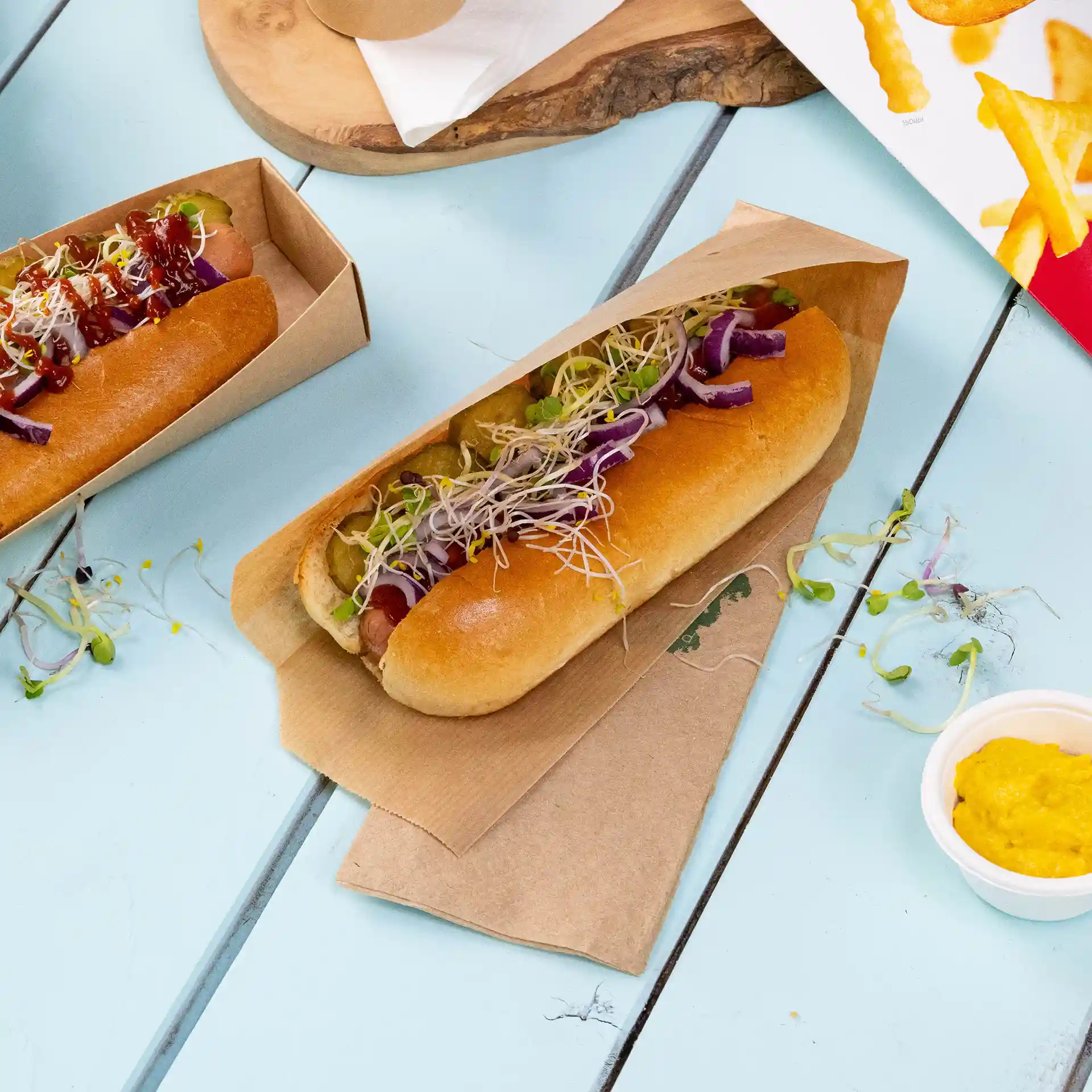 Dwa hot dogi w brązowych papierowych tackach, z różnymi warzywami i sosami, na jasnoniebieskiej powierzchni.