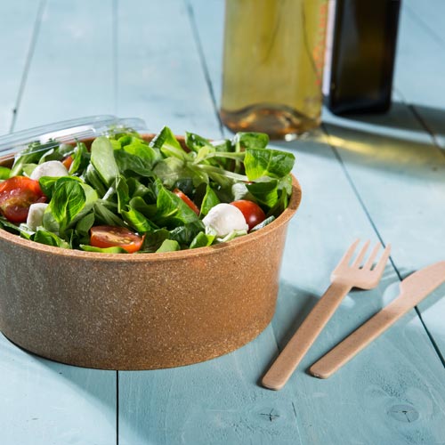 Brąbowy biodegradowalny miseczka wypełniona świeżą sałatą, z pomidorami i mozzarellą, obok drewnianych sztućców na niebieskiej drewnianej powierzchni.