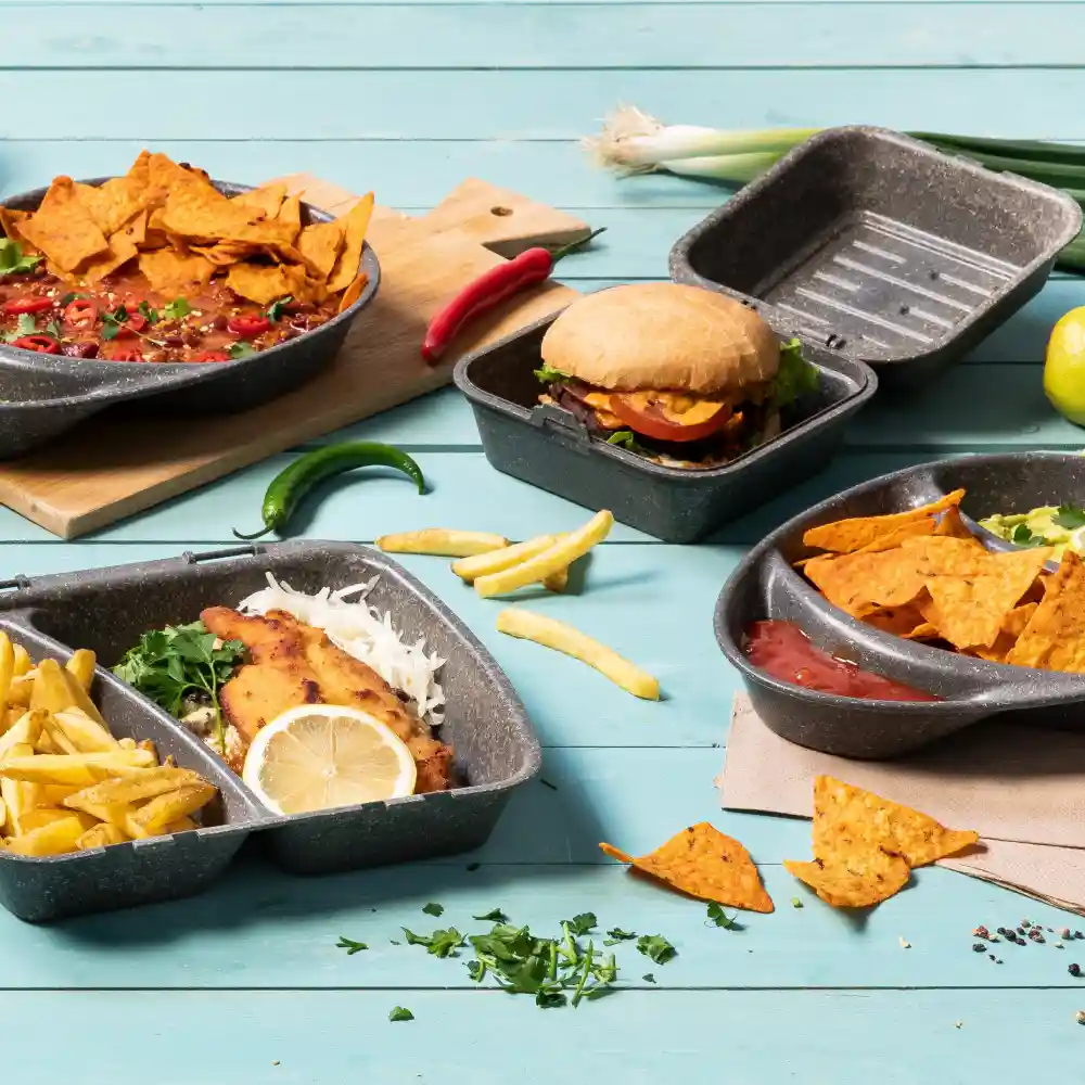 Różnorodność produktów spożywczych w wielokrotnego użytku pojemnikach, w tym nachosy, hamburger, frytki i ryba z cytryną, na niebieskiej powierzchni.