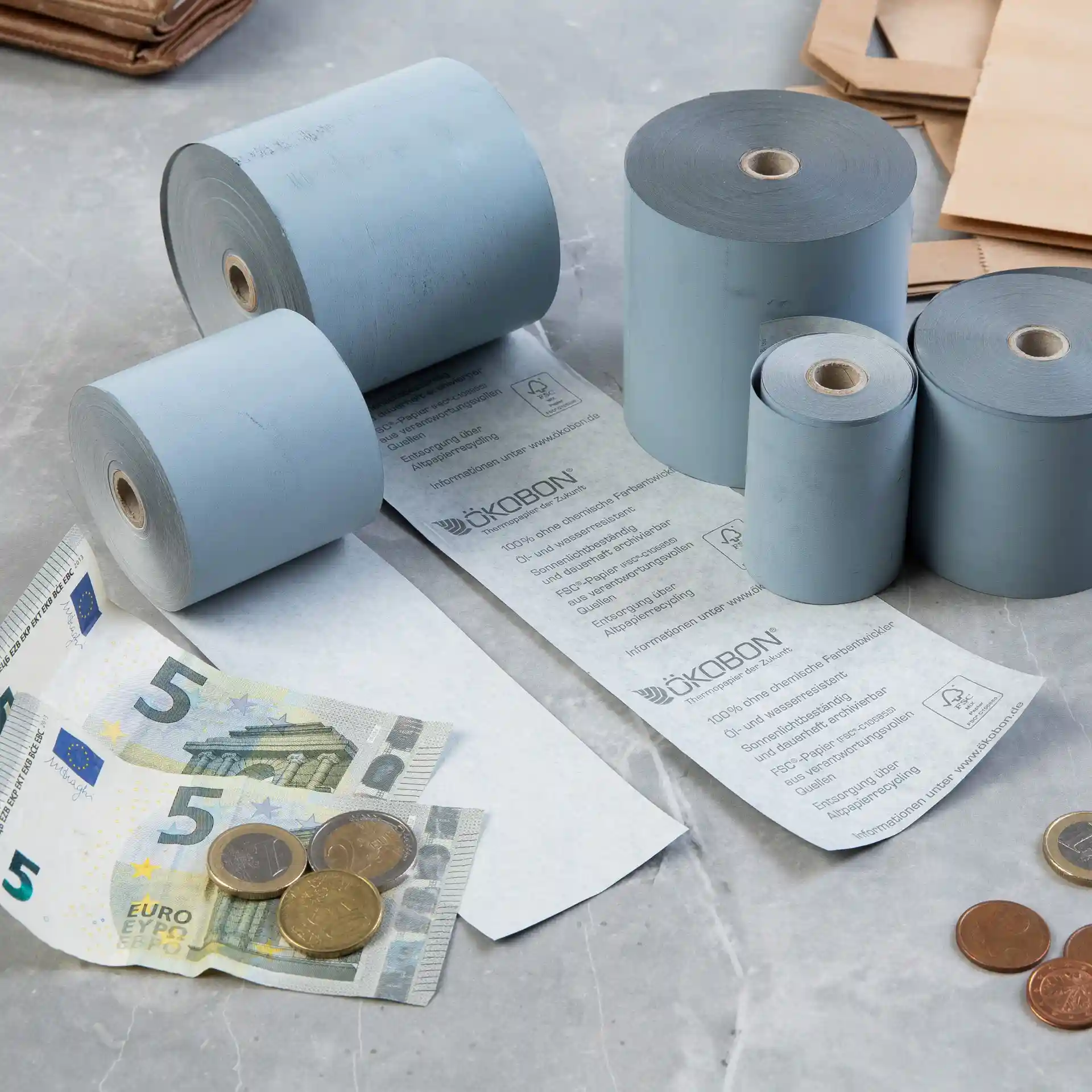 Obraz kilku szarych rolek papieru termicznego, banknotu 5 euro i monet na powierzchni.
