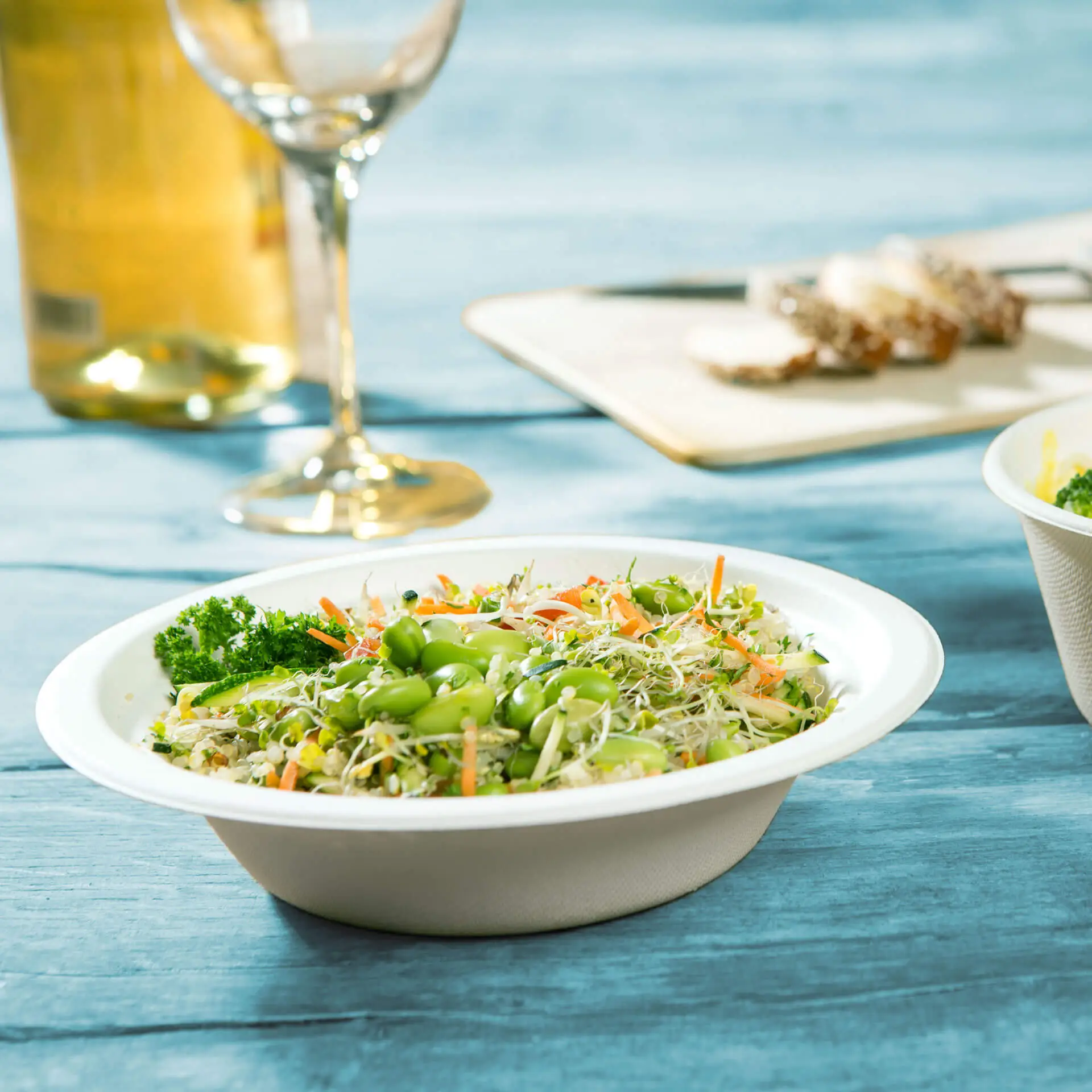 Miska wypełniona świeżą sałatką warzywną, w tym edamame i kiełkami, na niebieskim drewnianym stole, z kieliszkiem wina w tle.