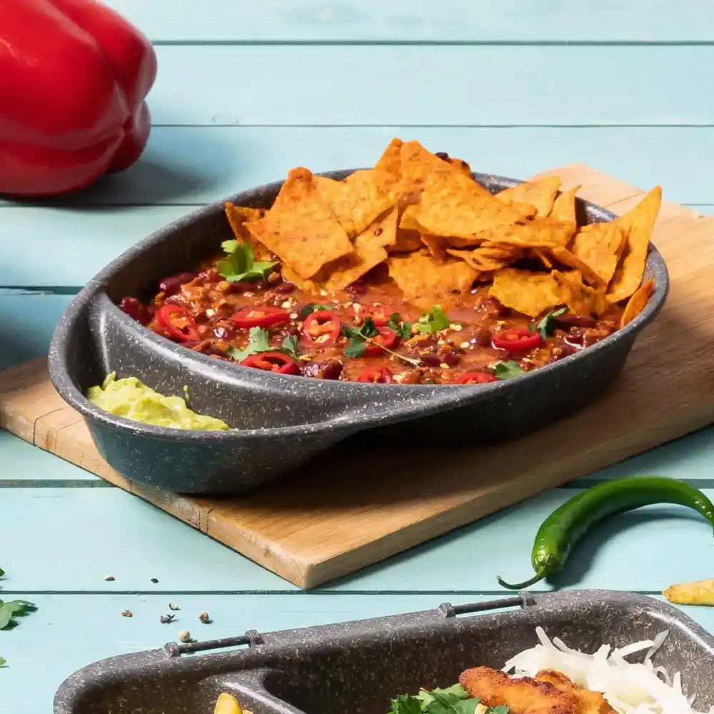 Miska z chili, polana chipsami tortilla, pokrojoną czerwoną papryką i kolendrą na drewnianej desce z dodatkami.